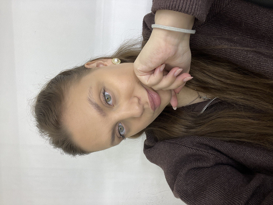 A young woman with light skin and green eyes poses for a selfie. She has brown hair, lightly styled, and is wearing a brown sweater with a visible silver necklace. A simple, silver bracelet is on her wrist, and a pearl earring is visible. Her hand is gently touching her face. The background is a plain white wall. The image is rotated sideways.