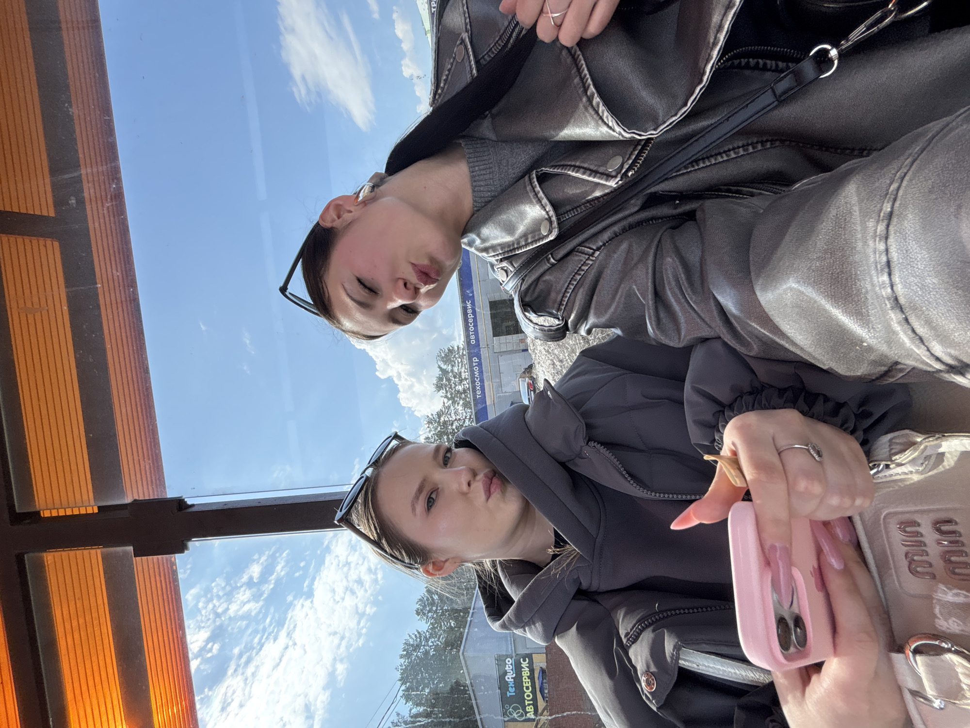 Two young women are captured in a selfie-style photograph, seemingly taken through a large window, possibly from a vehicle or building. Both are looking directly at the camera with a playful "duck face" expression.
The woman positioned on the left (when the image is correctly oriented) is wearing a dark gray hooded puffer jacket. She has sunglasses perched on her head and multiple rings on her fingers, including a golden, star-shaped or abstract ring and another with a small stone. Her fingernails are long and painted a light pink. She is holding a pink smartphone in a matching pink case in her left hand, and part of a light pink or beige handbag is visible, partially revealing the embossed text "MIU MIU".
The woman on the right is dressed in a shiny dark gray or metallic-colored faux leather jacket worn over a gray knitted sweater. She also wears sunglasses on her head and a silver-colored ring on her right ring finger.
The background visible through the window features a bright blue sky with scattered white clouds at the top. Below the sky, there are green trees and a commercial building. Text on the building in the background is visible in Cyrillic script: "Техосмотр" (Technical inspection) and "Автосервис" (Auto service), suggesting an automotive service center. On the far left of the window, there is a reflection or part of the interior structure with vertical orange-brown slats.
The lighting indicates a bright, sunny daytime setting. The overall mood of the image is casual and playful. The image is originally rotated 90 degrees counter-clockwise.