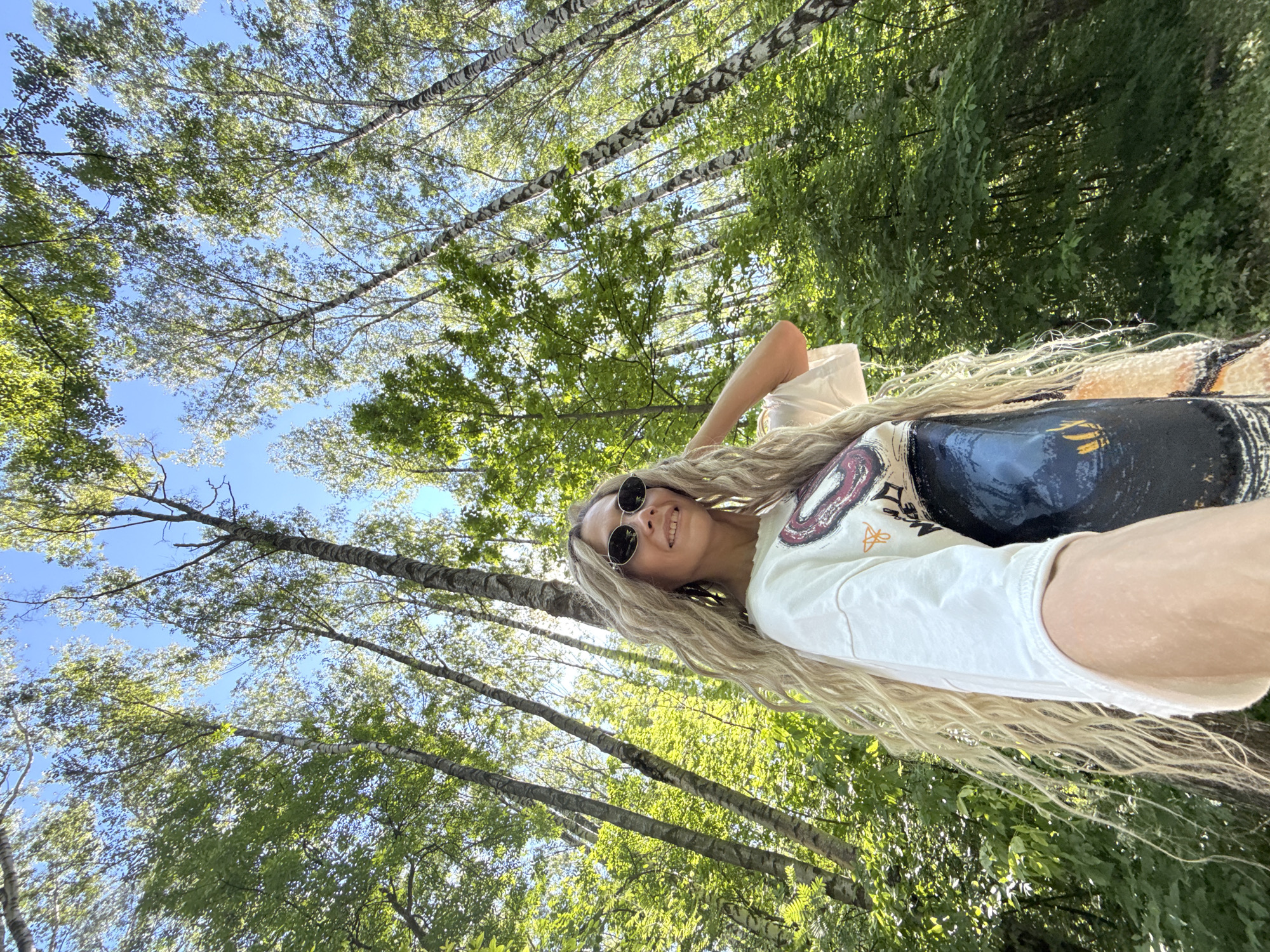 A young woman with long blonde hair and sunglasses smiles as she holds her hand up to her hair, standing in a forest in Rusanovo, Russia. The trees are tall and slender, with green leaves and white bark. The sun is shining through the trees, casting dappled shadows on the ground. The woman is wearing a white t-shirt with a graphic print and a dark skirt. She is standing at a low angle, looking up at the trees and the sky.