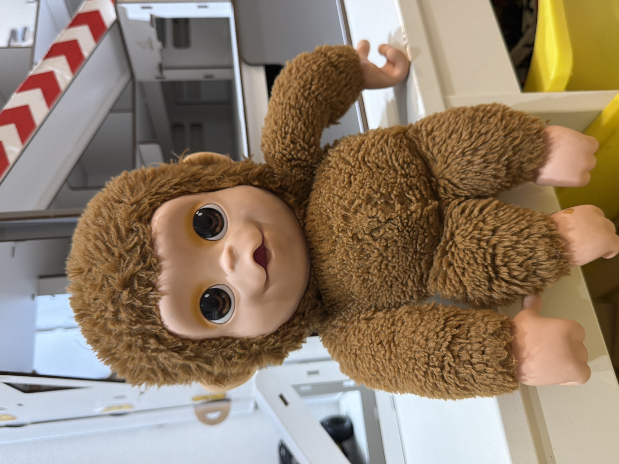 A plush monkey toy with a brown, shaggy fur body and a smooth, light-skinned face is the central subject of this image, which appears rotated 90 degrees clockwise. The monkey features large, dark brown eyes with prominent reflections, and its small mouth is slightly open, revealing a hint of a pink interior, giving it a gentle and curious expression. Its hands and feet are made of a smooth, peach-colored plastic, offering a textural contrast to its furry body. The monkey lies on its back on a light-colored surface, possibly a shelf or counter.
The background reveals an indoor setting, characterized by geometric shapes and neutral tones. To the upper left of the monkey's head (in the rotated view), there's a white structure with bold red and white diagonal stripes, resembling a barrier, ramp, or a safety marker. Beyond this, a series of white and off-white panels or shelves are visible, creating a structured, possibly modern, interior environment. On the far right, a vibrant yellow object or surface adds a pop of color to the scene. There is no specific activity taking place, as the monkey is a stationary toy. The lighting is consistent with an indoor setting, likely artificial or ambient daylight. No visible text can be discerned in the image. This scene is set in Sofiivska Borshchahivka, Ukraine.
