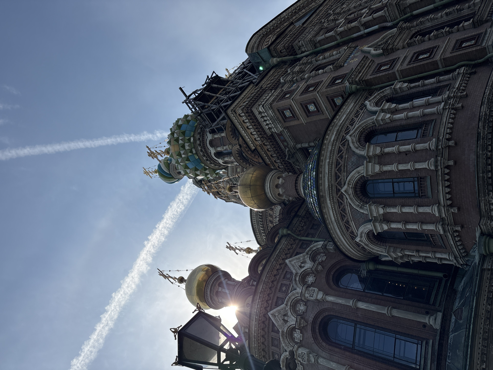 The image is a low-angle shot of the Church of the Savior on Spilled Blood in Saint Petersburg, Russia, against a bright, sunny sky with two prominent contrails. The church's ornate facade is visible, with its domes, arched windows, and intricate brickwork. Some scaffolding is present on one of the domes, indicating ongoing restoration or maintenance. A street lamp is visible in the foreground, partially obscuring the base of the church. The sun is positioned behind one of the domes, causing a strong lens flare.