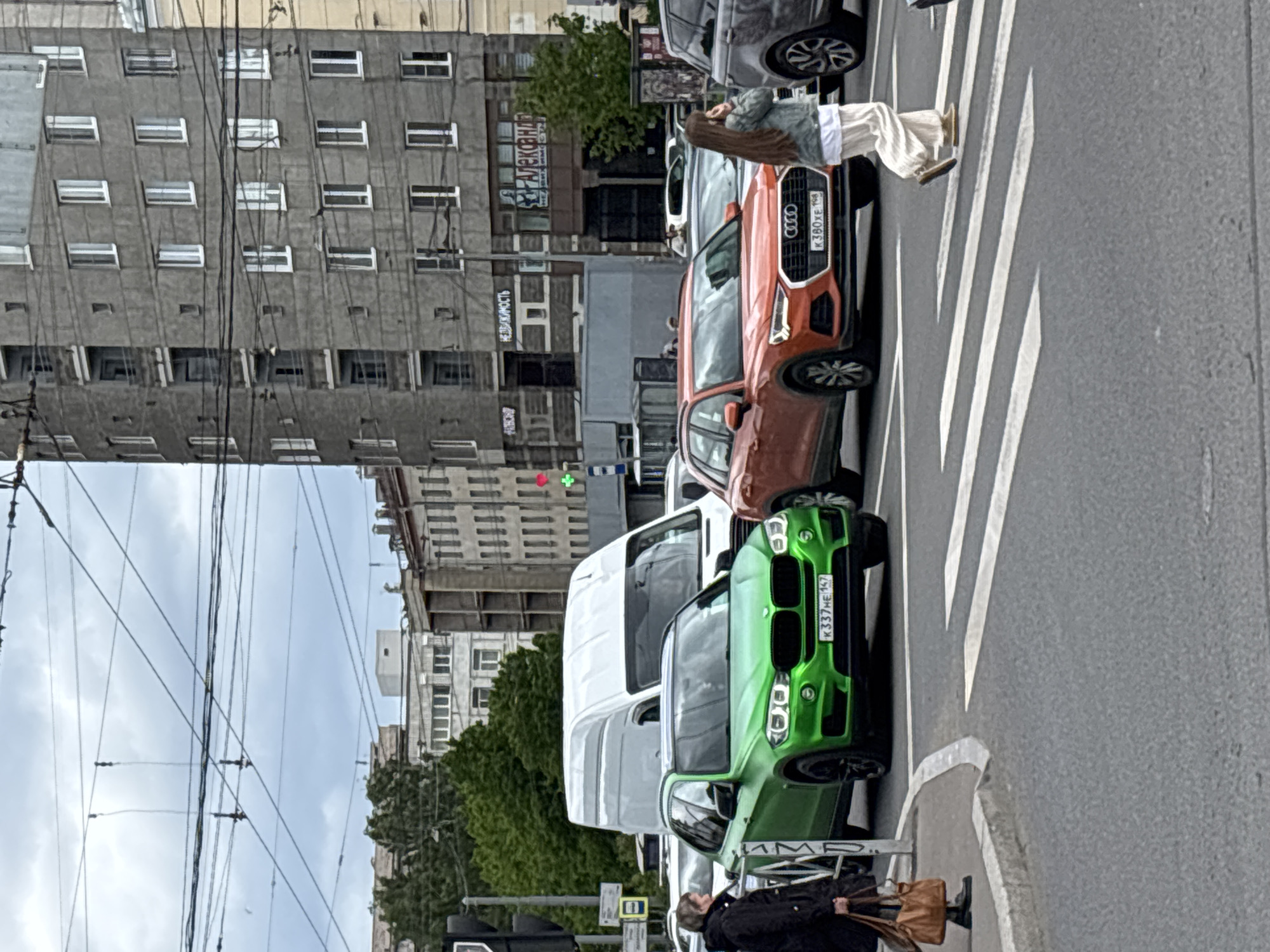A photograph captures a street scene in Saint Petersburg, Russia, featuring several vehicles stopped at a crosswalk. A bright green BMW SUV with the license plate "K 337 HE 197" is positioned closest to the camera. An orange Audi SUV with the license plate "K 380 XE 198" can be seen in front of it, as well as a white van. Pedestrians are present: one female is in a white skirt and a light jacket is walking at the crosswalk. Another pedestrian is standing on the sidewalk in front of the green car. The buildings in the background are typical of Saint Petersburg's urban architecture. Overhead electrical wires are visible against the sky. Street signs, traffic lights with a red light indicating stop, and advertisements are also observable in the scene.