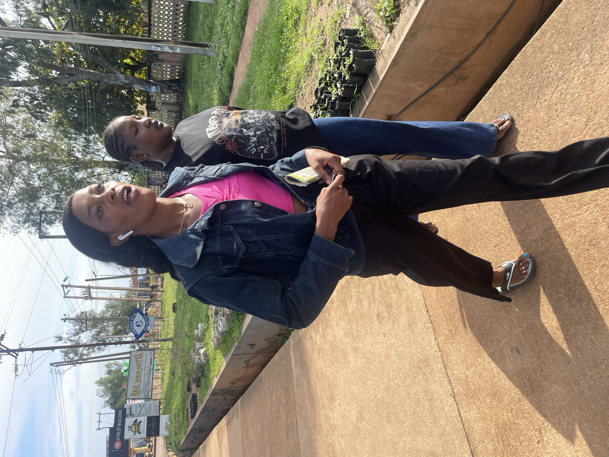 In Jos, Nigeria, two young women walk along a concrete sidewalk. The woman in the foreground wears a denim jacket over a bright pink crop top and black pants, with white earbuds in her ears. She holds a folded pamphlet or paper in her right hand. The woman behind her wears a black t-shirt with a graphic design and blue jeans. They are walking in the same direction. In the background, there are signs for "18 STREET BUILDING" and "RAIL VIEW HOTEL", as well as a blue sign with a "JORDAN EYE HOSPITAL" logo. Electrical poles and trees are visible, and a grassy area with rows of small potted plants is to their right. The scene appears to be outdoors during daylight.