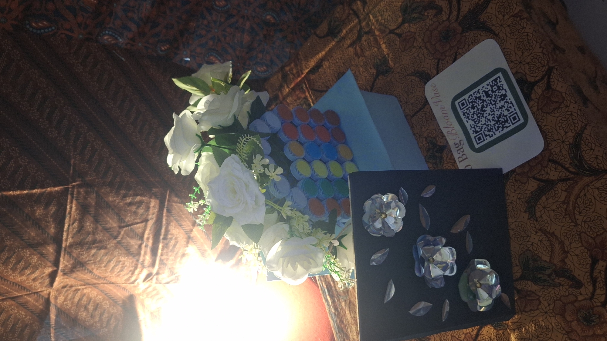 The image displays an arrangement of decorative items on a patterned fabric surface, likely a table or cloth, consistent with traditional Indonesian batik patterns in shades of brown, orange, and dark brown. A strong light source from the lower left illuminates the scene, casting shadows and creating glare.
The primary subjects include a bouquet of artificial white roses with green foliage, positioned centrally. To its right, a light blue box or container is visible, adorned with a grid of colorful, circular objects, possibly bottle caps, arranged in rows of red, blue, green, yellow, and orange. Below this box, and to the right of the flowers, lies a black rectangular object decorated with three iridescent, faceted flower-shaped embellishments and several smaller, leaf-like iridescent pieces.
In the upper right corner, a white card with rounded corners is placed, featuring a prominent QR code. Visible text on this card reads "Bag Bloom Vase," with the initial part of "Bag" slightly obscured.
The scene appears to be an indoor setup, possibly showcasing gifts or handcrafted items. No human figures are present, and no direct activity is taking place beyond the static arrangement of the objects. The overall impression is one of presentation or display.