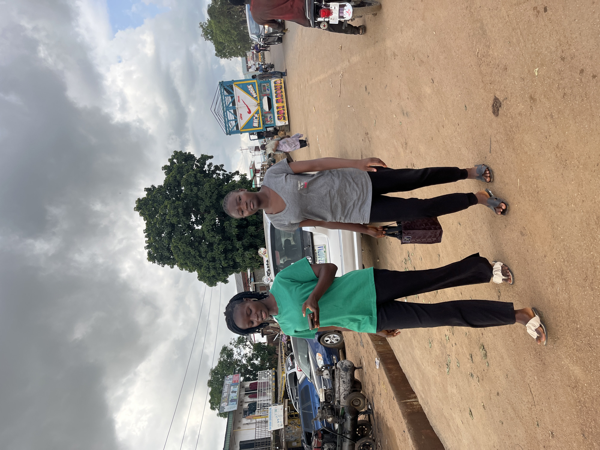 In Rafin Par, Nigeria, two young women stand on a dusty, unpaved road under a cloudy sky. The woman in front, wearing a bright green t-shirt and black trousers, holds a mobile phone. She is looking down with her eyes closed. Behind her, another young woman in a grey t-shirt and black trousers stands with her arms crossed and a slight smile on her face. She carries a dark handbag.
The scene is a roadside with various vehicles, including a white car, a blue car, and a motorcycle visible in the background. A distinctive blue and white truck with the words "WORK" and "NICE" printed on it is parked nearby. There are also buildings and structures suggestive of a market or commercial area. The presence of electrical wires and trees indicates an outdoor, possibly urban or semi-urban, setting. The weather appears overcast, suggesting a possible time of day when the sun is not directly visible. The overall impression is of a casual, everyday moment captured on a street in Nigeria.