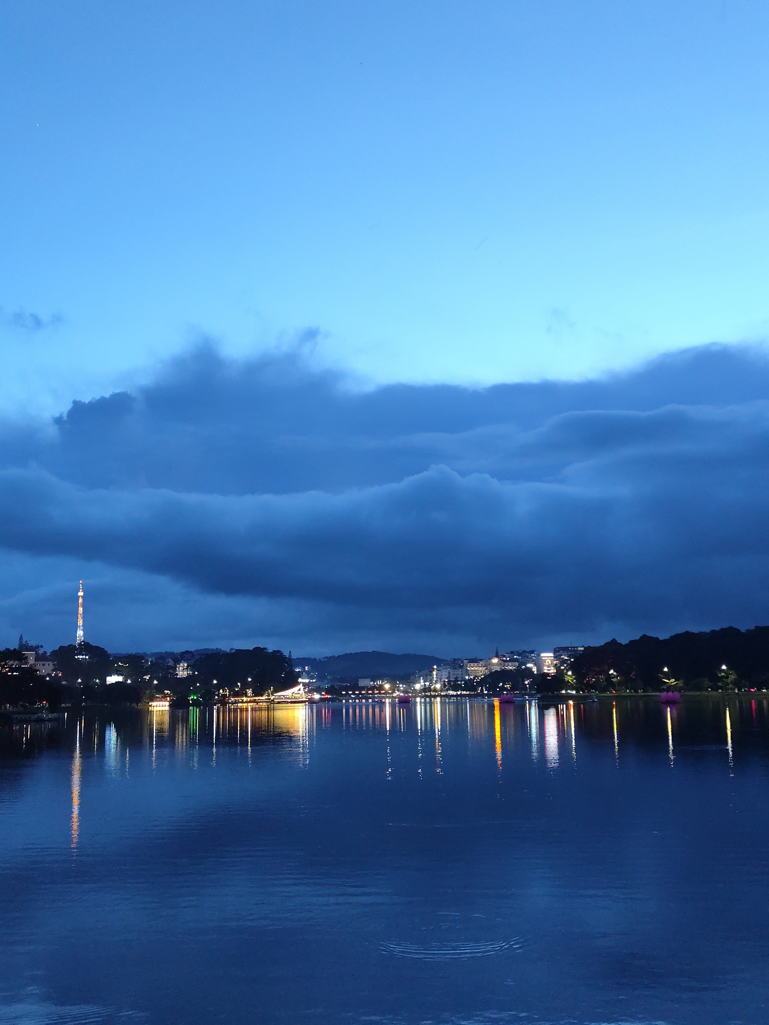 The image depicts a nighttime scene at Xuan Huong Lake in Da Lat, Vietnam. The foreground is dominated by the dark, still surface of the lake reflecting the city lights. The city skyline is visible in the mid-ground, illuminated with various lights, including a prominent communications tower. The background features heavy, dark clouds that fill the sky, with a lighter, blue sky visible above. The water reflects the light producing a ripple effect.
