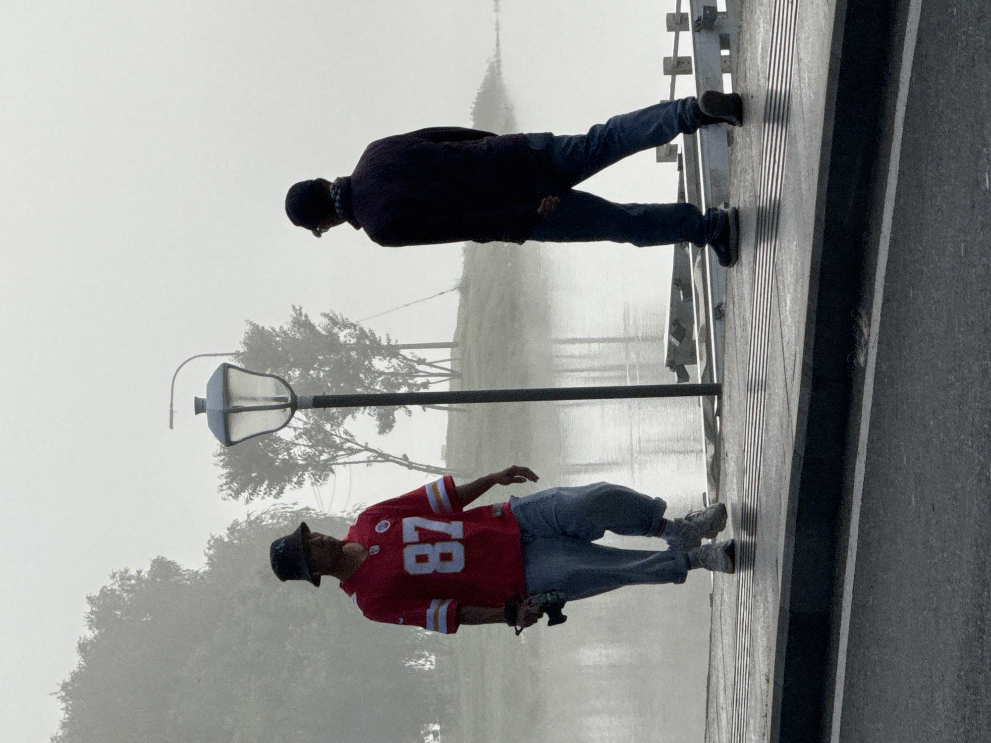Two individuals are walking on a pier in Da Lat, Vietnam, under a dense fog. The person on the left, facing the camera, is wearing a red jersey with the number "87" and a dark bucket hat. They are holding a camera in their right hand and are dressed in baggy blue jeans and sneakers. The person on the right is walking away from the camera, wearing a dark jacket, blue jeans, and a baseball cap. They have a scarf or bandana around their neck. In the background, trees are visible through the fog, and a lamppost stands between the two individuals. The pier is made of wood with metal railing structures. The foreground shows a wet asphalt road with white lines. The foggy atmosphere creates a diffused, hazy environment, obscuring details in the distance and creating a sense of isolation.