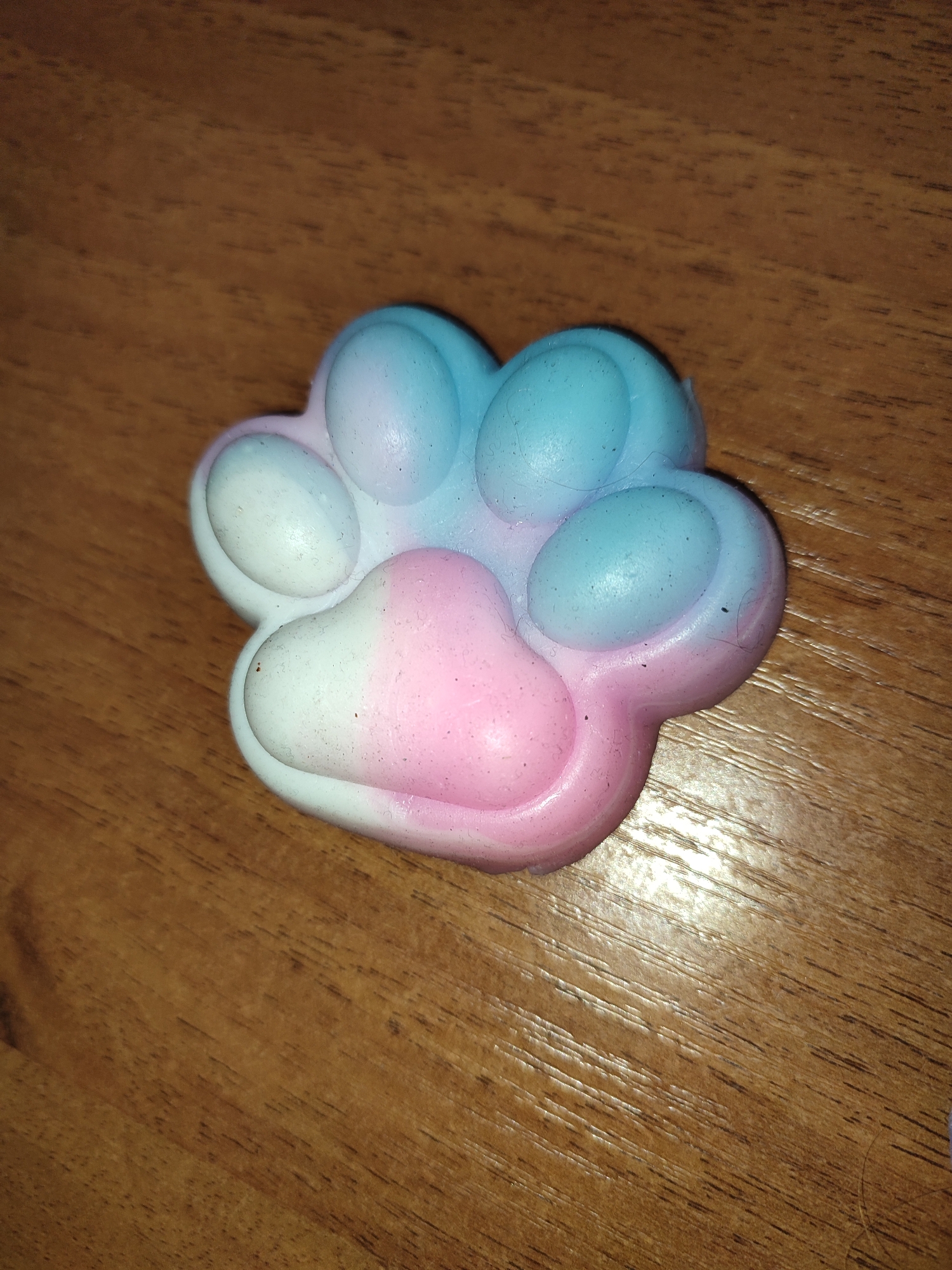 A paw-shaped pop-it fidget toy lies on a wooden surface. The toy is a pastel gradient of pink, white, and blue, with five circular bubbles. The wooden surface has a visible grain. There is no indication of the location in the city of Shyshaky, Ukraine.