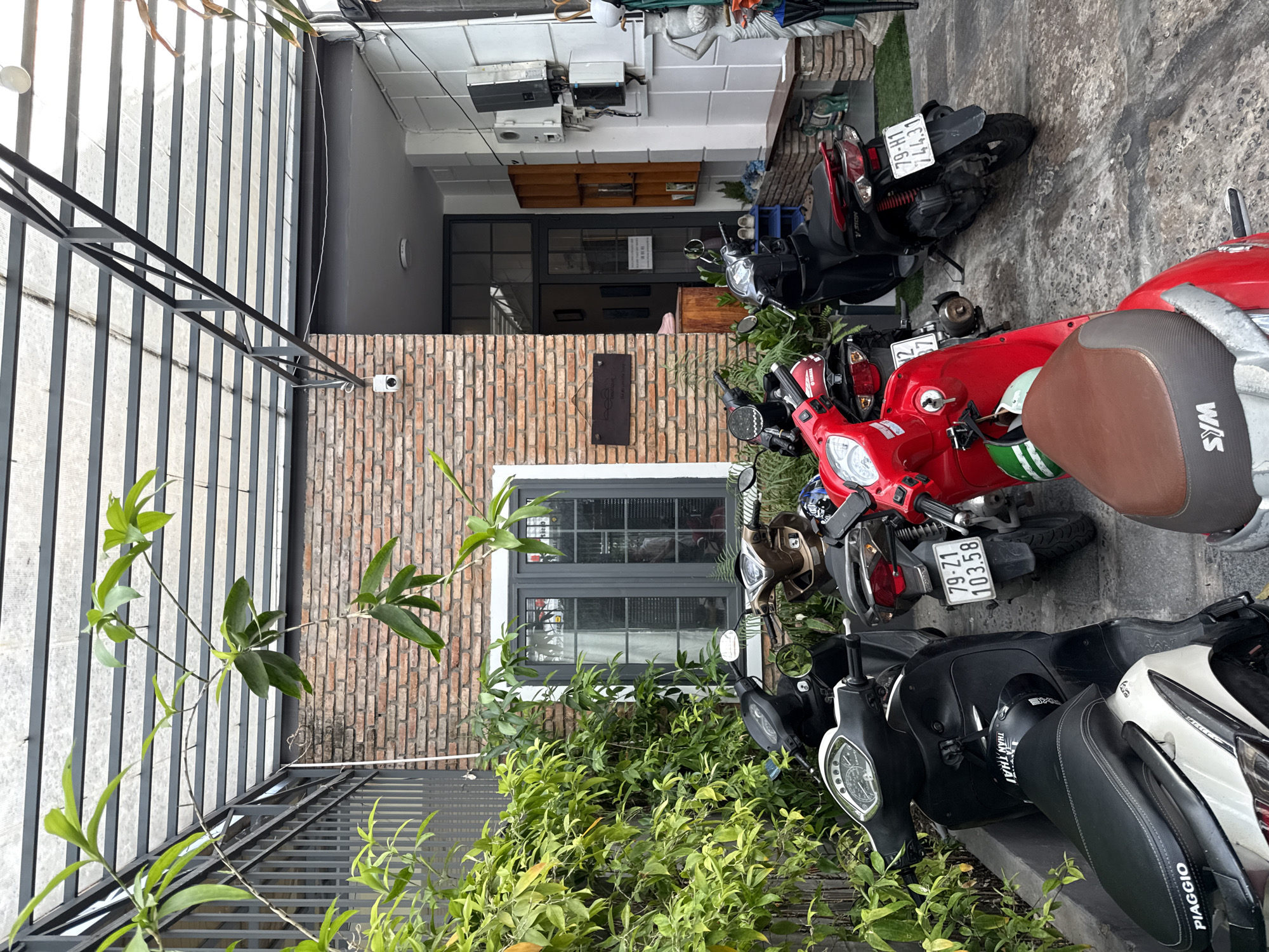 The image depicts an outdoor scene in Nha Trang, Vietnam, likely a residential or commercial establishment. A brick building with a grey-framed window forms the background. In front of the building, several scooters are parked on a concrete surface. Lush green foliage is visible, with plants growing along the left side and trailing from an overhead structure. A security camera is mounted on the brick wall near the entrance. The overall atmosphere suggests a quiet, possibly overcast day.