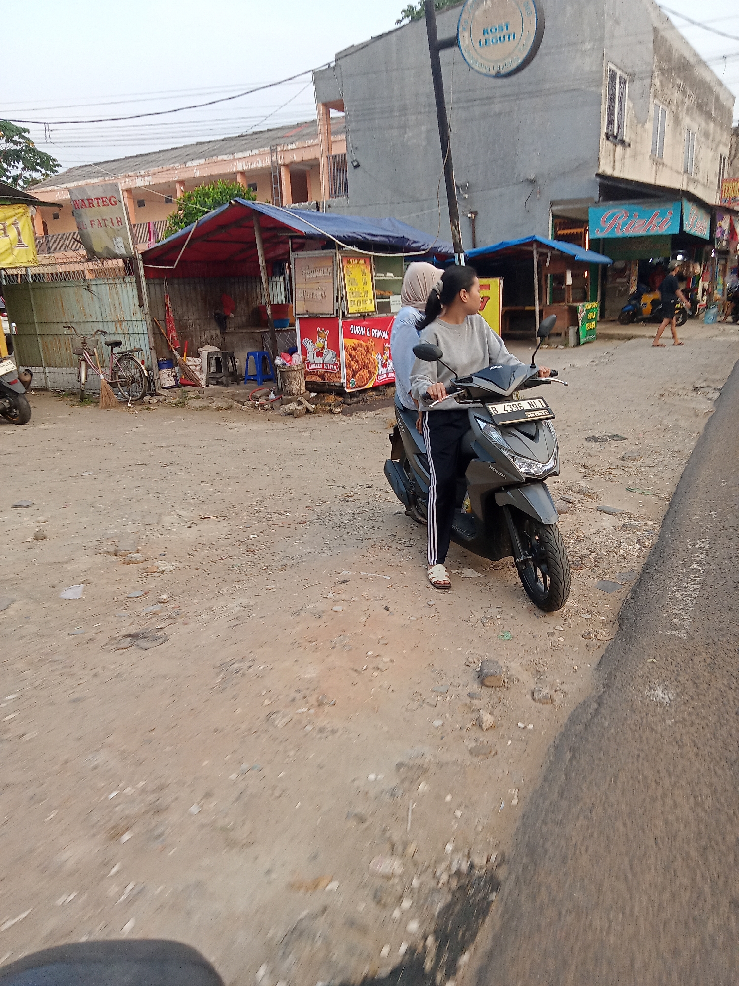 In an outdoor scene in South Tangerang, Indonesia, two individuals are positioned on a dark gray Honda Beat scooter, which is stopped on a dirt and gravel roadside. The scooter's license plate reads "B 4396 HLT". The individual in the foreground, wearing a gray sweatshirt and black pants with white stripes, is operating the scooter. They have on white sandals and are looking to their right. Behind them, a second individual, wearing a light blue hijab and a light blue shirt, is seated on the scooter.
The background features a collection of roadside food stalls and buildings. To the left, a stall with a red and blue awning displays a banner that says "WARTEG FATIH". Another stall, further back, has a yellow sign. To the right of the scooter, a building with a gray facade has a blue sign with the word "RIZKI" in cursive. Further down the street, another person is visible walking. The environment suggests a commercial area with informal businesses. Power lines are visible overhead.