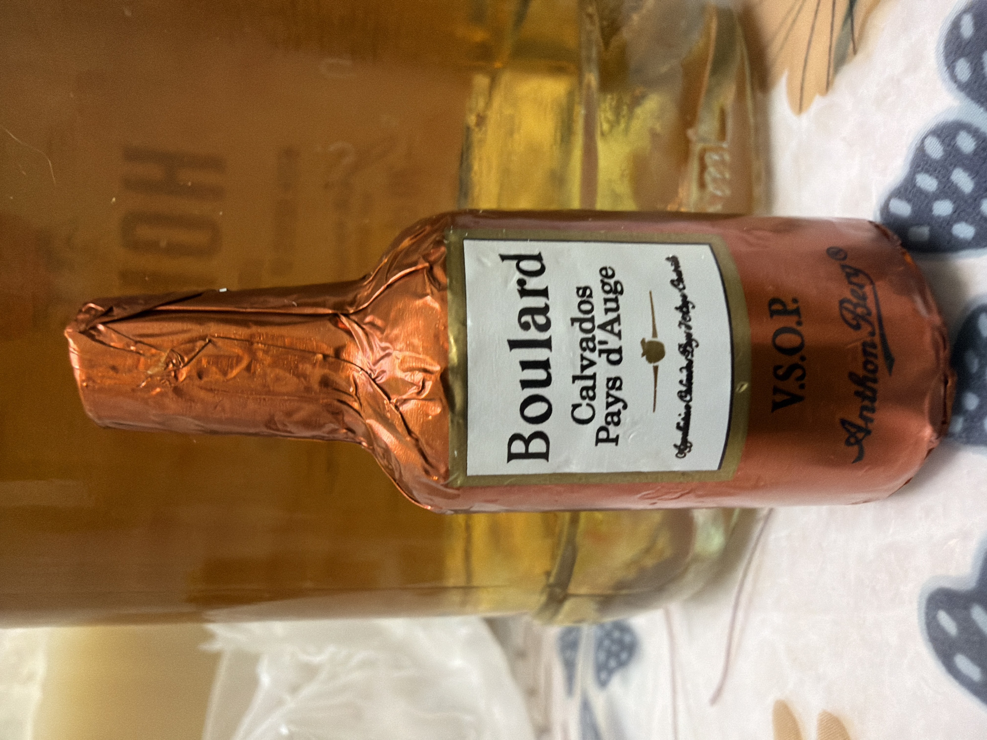 A small, foil-wrapped bottle of Boulard Calvados Pays d'Auge V.S.O.P. liqueur is the central subject of the image. The bottle is wrapped in crinkled, metallic copper-colored foil. A white label is visible on the front of the bottle, featuring black text that reads "Boulard Calvados Pays d'Auge" and "V.S.O.P." Below this, in smaller cursive text, is "Authentic Calvados Pays d'Auge." The bottom of the foil is partially unwrapped, revealing a dark brown color. Underneath the label, the foil has "Anthon Berg" printed in a cursive font. The bottle is positioned in front of a much larger, blurry glass bottle or container filled with a golden-brown liquid, possibly more liqueur or another beverage. The background is out of focus, but appears to be a table covered with a patterned cloth, with some indistinct objects and shadows. The lighting suggests an indoor setting, possibly with ambient light from a window. There are no people or animals visible in the image. The scene does not provide any specific information about its location in the city of Kyiv, Ukraine.