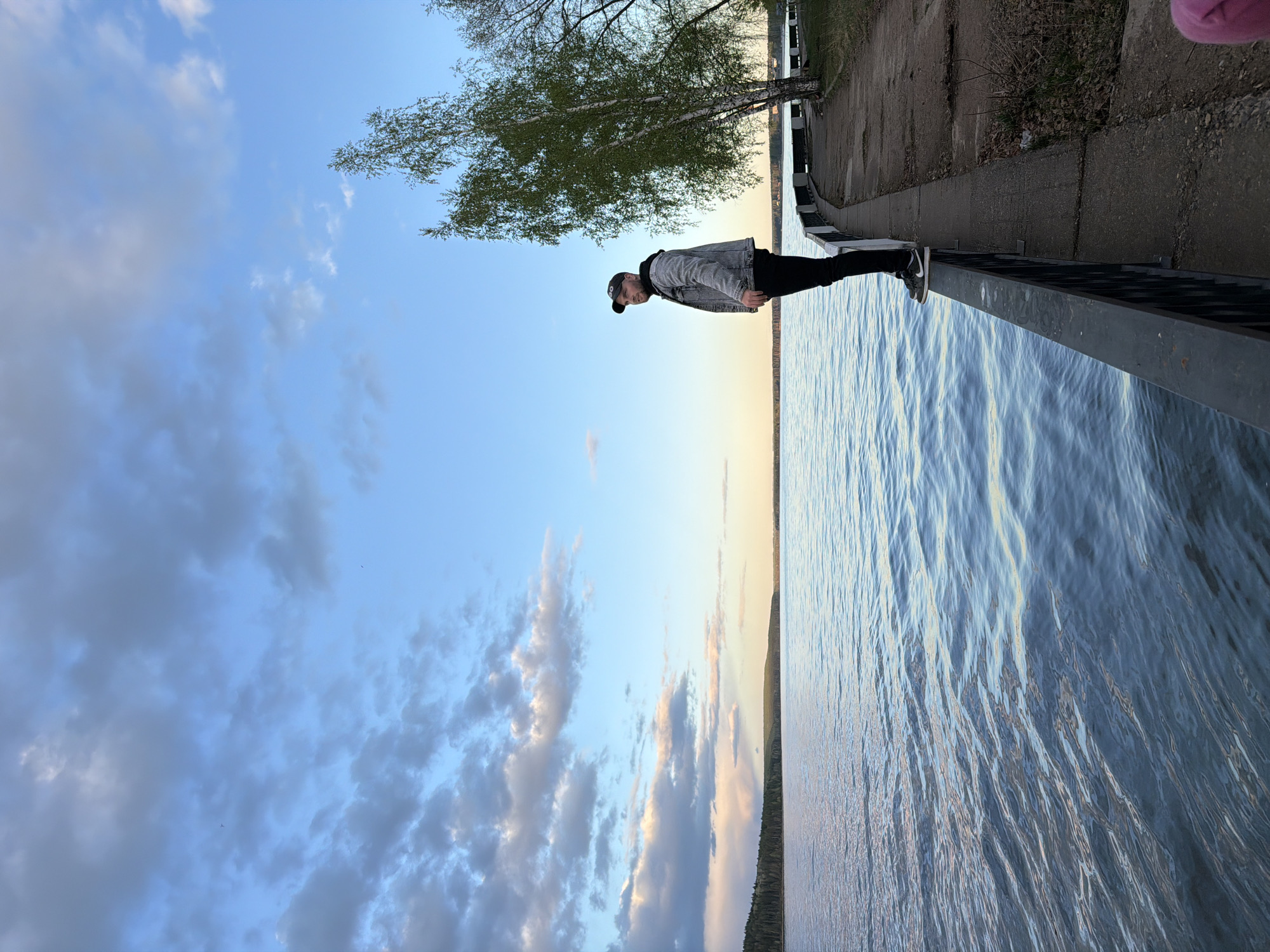 A person stands on a concrete railing bordering a body of water beneath a partly cloudy sky. The location is likely in or near the city of Chaikovsky, Russia. The individual is wearing dark pants, a gray jacket, and a baseball cap. A tree with light-colored bark is visible near the person. The body of water reflects light, and a concrete walkway or embankment is situated alongside the railing.