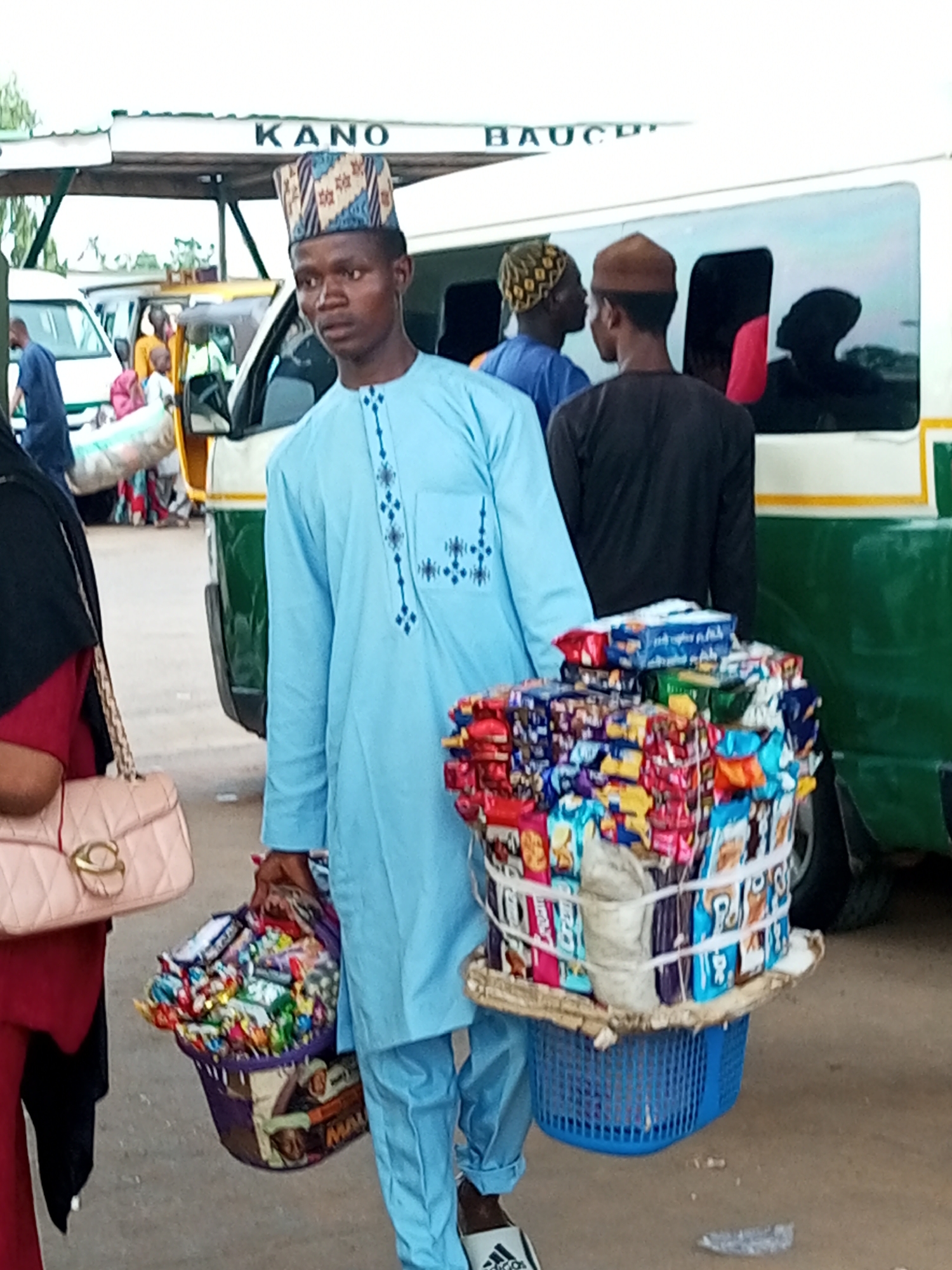 In Gombe, Nigeria, a young man wearing a light blue traditional outfit and a patterned cap stands in front of a white bus with green accents, holding a large basket of snacks. The bus has "KANO - BAUCHI" printed on its side, indicating it is a transport vehicle traveling between these cities. The man is carrying the basket of snacks, which are individually packaged items, on his shoulder. To his left, a woman in a red top and black headscarf holds a light pink handbag with a gold clasp. Other people are visible in the background near other vehicles. The scene appears to be at a bus station or transport hub.