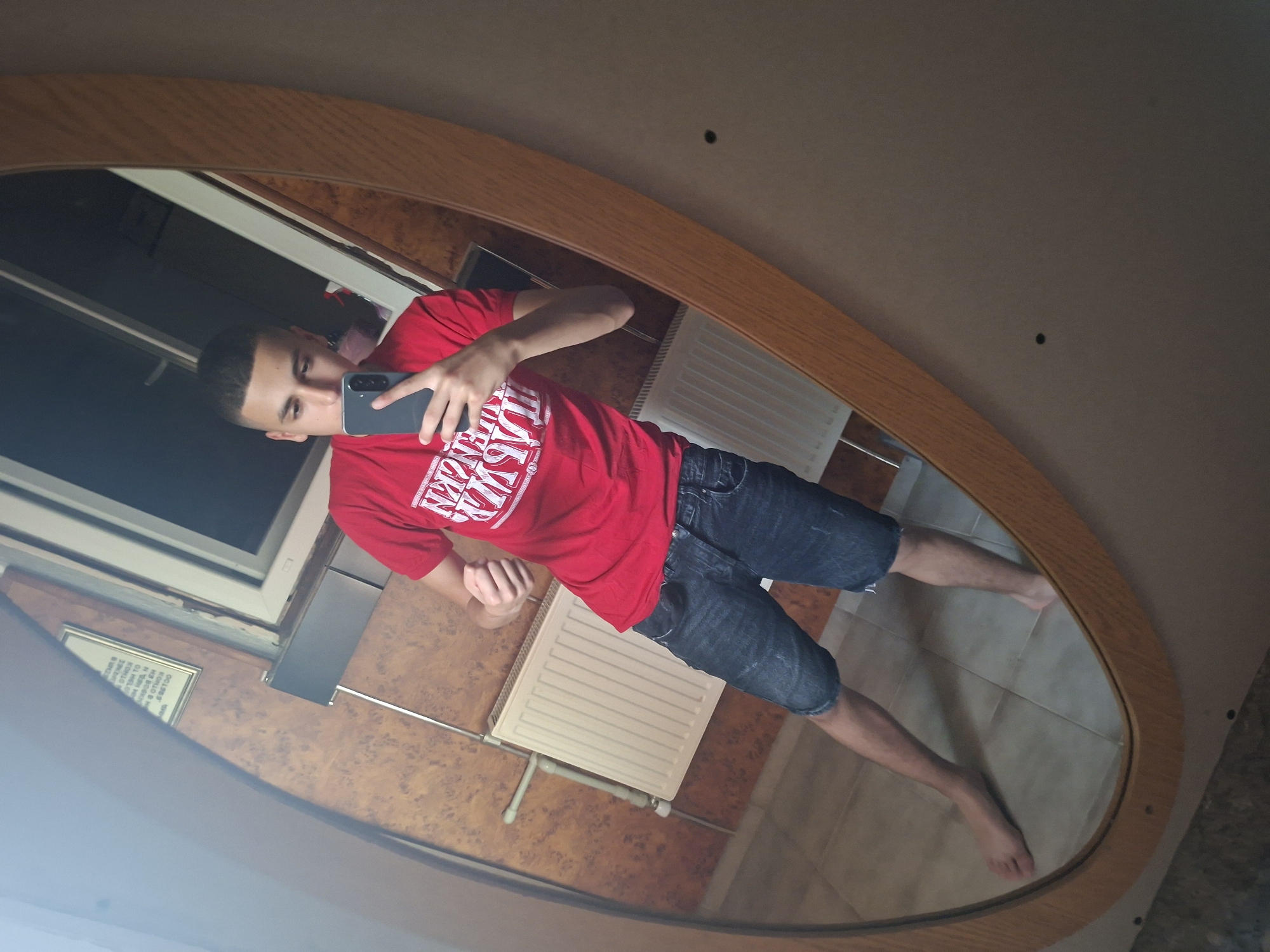 This media file features a mirror selfie of a young man taken indoors. The man, who appears to be in his late teens or early twenties, has short, dark hair and is clean-shaven. He is wearing a red t-shirt with white text that partially reads "OLD WEST" and dark denim shorts, and he is barefoot. He holds a dark-colored smartphone in his right hand, positioned to capture his reflection. His left arm is bent, with his hand forming a fist.
The reflection is captured in a large, irregularly shaped mirror with a light wooden frame. The background visible in the mirror includes a window with a white frame in the upper left, suggesting daytime lighting. A white radiator is mounted on the wall behind the man's lower body, and a framed item with illegible text is partially visible on the wall to the left of the radiator. The floor, visible beneath the man's feet, is tiled in a light brown or beige color. The upper part of the reflection shows a plain, light brown ceiling or wall. The man's expression is neutral as he looks towards the phone. The setting appears to be a functional interior space in a building located in Basarbovo, Bulgaria.