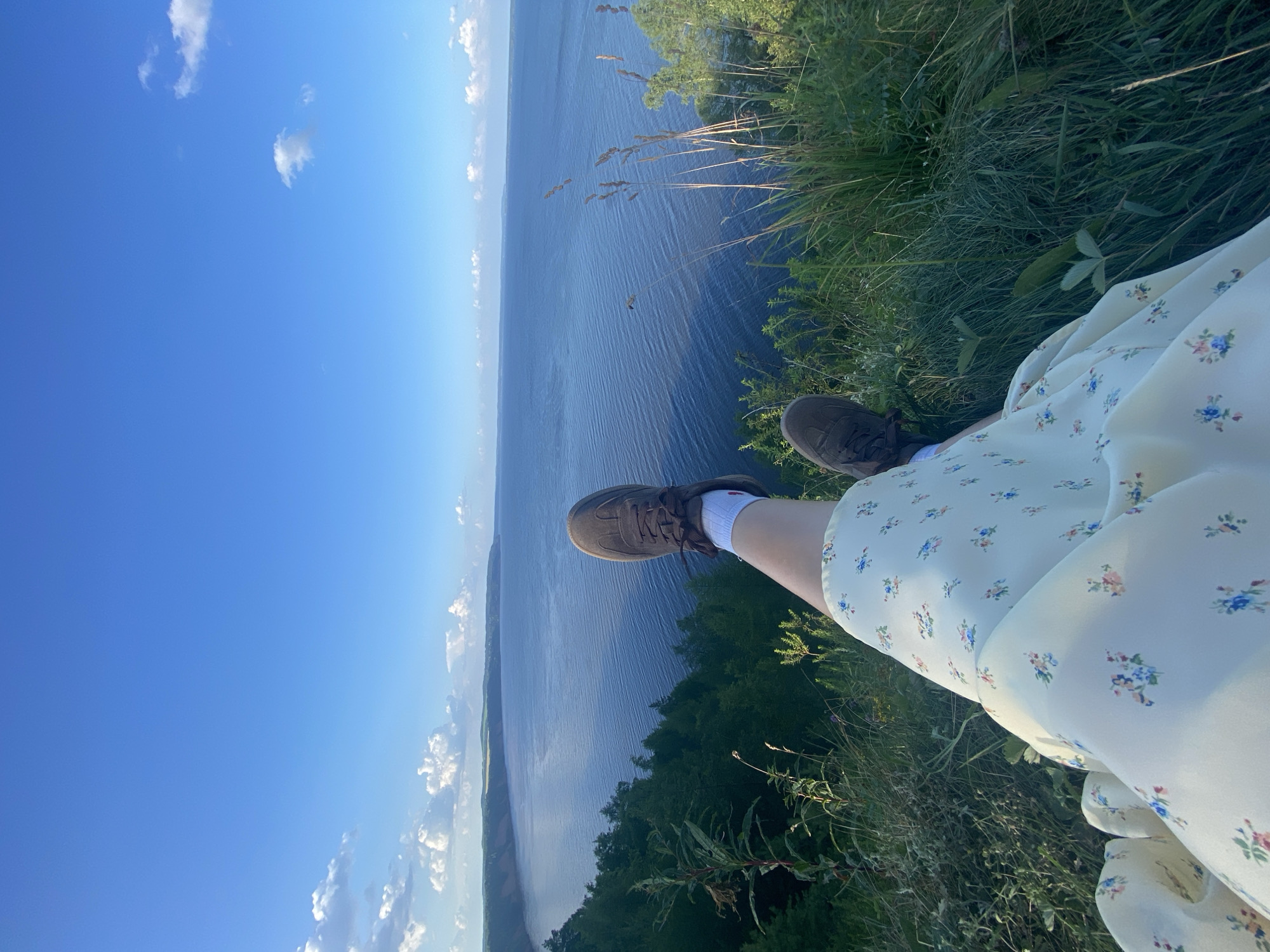 The image shows a person's legs and feet, clad in brown sneakers and white socks, resting on a grassy cliff overlooking a wide body of water. The person is wearing a light-colored, floral-patterned dress. The landscape is dominated by a vast blue sky with scattered white clouds. Below the cliff, a dense green forest lines the shore, which curves into the distance. The water, likely a river or a bay, appears calm with subtle ripples. The overall scene conveys a sense of peaceful contemplation and appreciation of nature. The specific location of Kostovaty, Russia, is mentioned in the prompt, implying this scene is situated there.