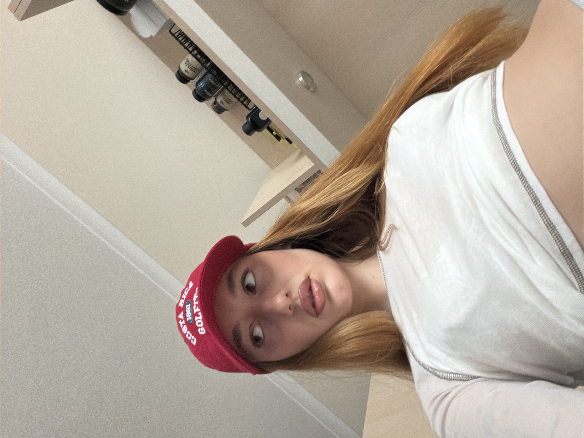 A young woman with long, strawberry blonde hair is wearing a red "Costa Rica Golf 1981" cap and a white long-sleeved top. She is looking at the camera with a slight pout. The background shows an off-white wall with shelves containing various bottles. The location is not discernible as Sofia, Bulgaria without additional context or identifiable landmarks.