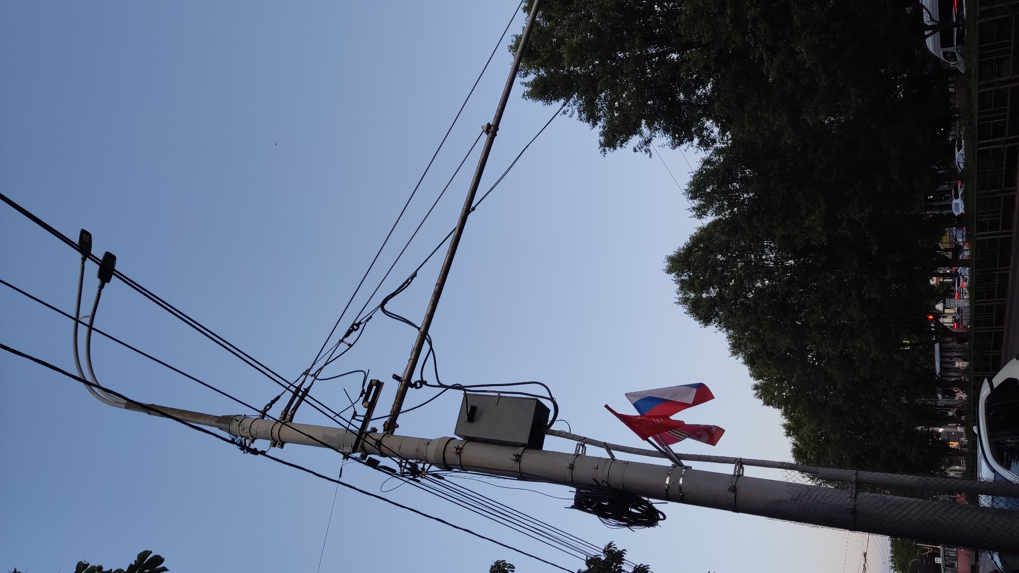 The image depicts a street scene in Krasnodar, Russia, dominated by a utility pole with numerous wires and two flags attached. The pole, situated on the left side of the frame, is a prominent feature, supporting a gray electrical box and a complex network of overhead wires. Two small flags are affixed to the pole: one appears to be the flag of Russia, with its white, blue, and red horizontal stripes, and the other is a red flag with a golden emblem, possibly representing a specific organization or event.
In the background, a row of lush, dark green trees lines the street, suggesting a park or boulevard. Beyond the trees, a street with traffic is visible, including cars and what appears to be a tram or trolleybus. The sky is a clear, pale blue, indicating it is either early morning or late evening. The overall impression is that of a typical urban street in a Russian city, with a focus on the infrastructure and a touch of patriotic or ceremonial display with the flags. There are no discernible people in clear focus or performing any specific actions.