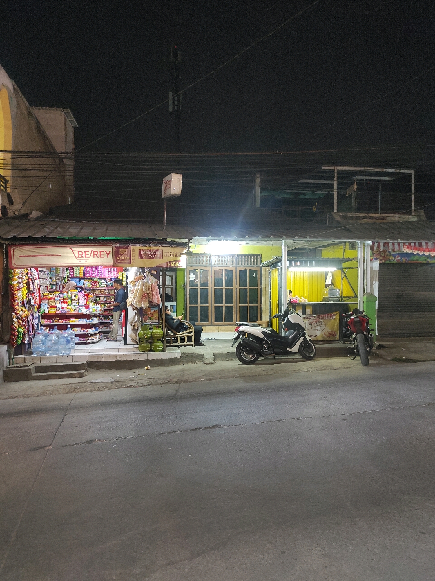 This is a night-time outdoor shot of a small convenience store and a food stall in Padalarang, Indonesia. The convenience store, labeled "RE/REY," is brightly lit and filled with various products displayed on shelves. Two men are visible near the store: one is standing and looking at the products, while another is seated on a chair outside the store, appearing to be using a mobile phone. Several large bottles of water are placed on the steps leading up to the store.
Adjacent to the convenience store is a food stall with yellow walls, also illuminated by lights. A white scooter and a red motorcycle are parked in front of the stall. Another person, also seated on a chair, is visible behind the counter of the food stall. The street in front of the stores appears to be made of asphalt, with some cracks and debris visible. A tall telecommunications tower is present in the background, and power lines are strung across the scene. The overall impression is of a typical street scene in a residential or commercial area at night.