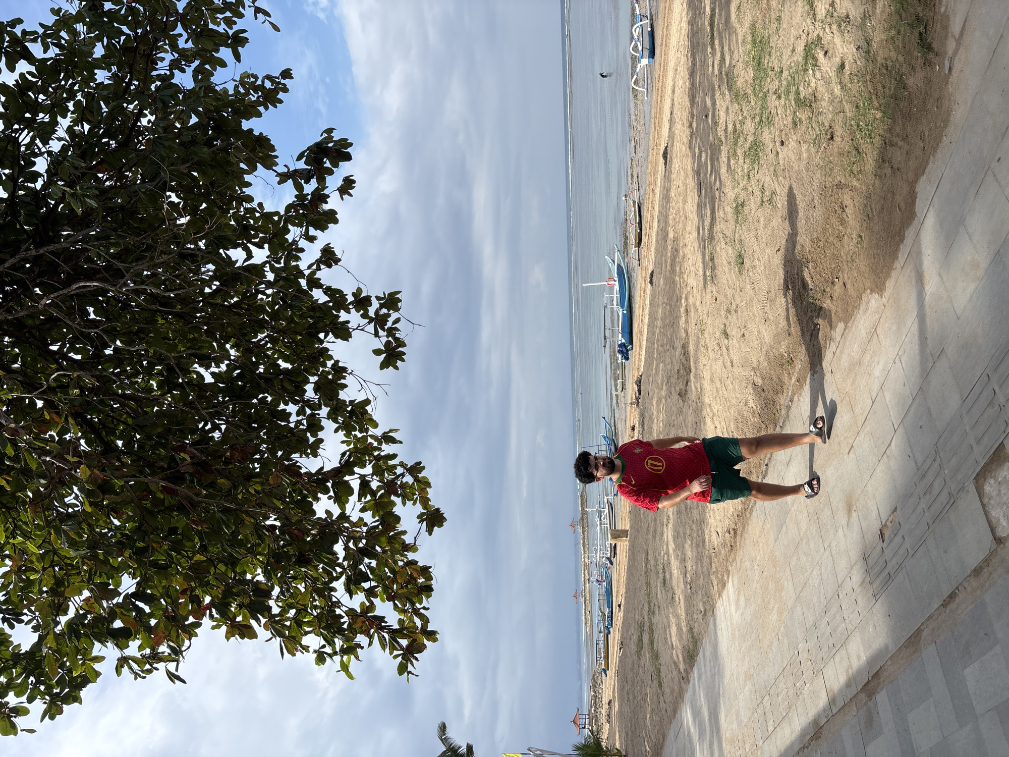 A man wearing a red shirt with a circular logo and green shorts walks on a paved pathway next to a sandy beach in Denpasar, Indonesia. The beach has several small boats pulled ashore and in the shallow water. The ocean stretches to the horizon under a partly cloudy sky. Lush green tree branches hang over the scene from the top of the frame, casting shadows on the pathway and beach.