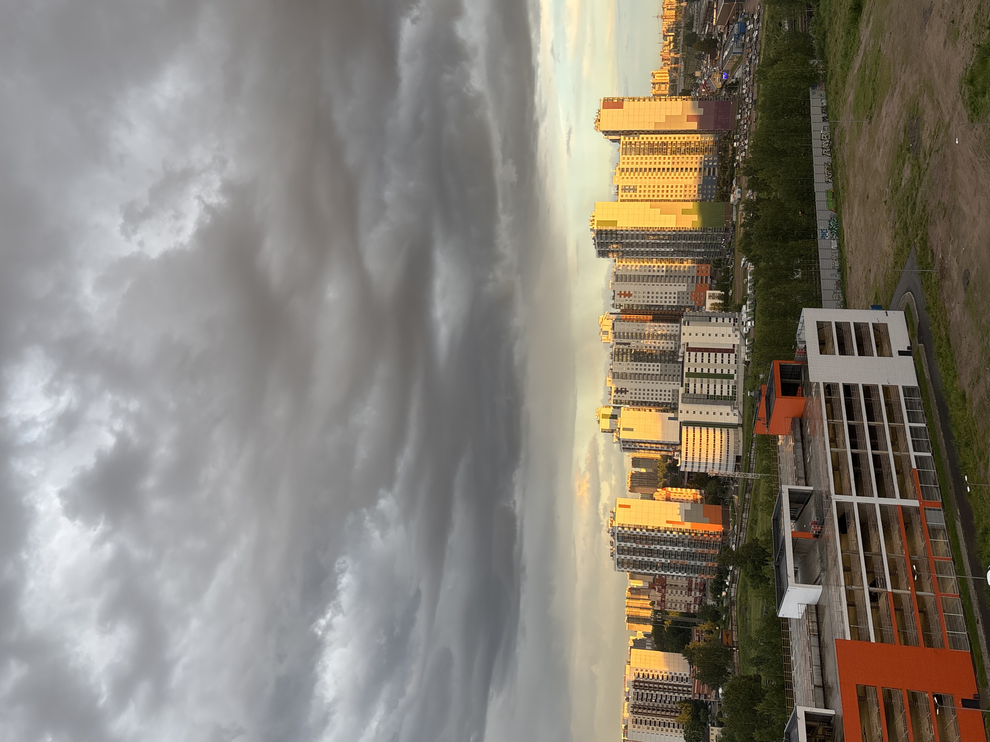 This is a vertical, eye-level, medium shot of a city skyline under a dramatic sky. The left half of the image is dominated by dark, heavy storm clouds, creating a sense of foreboding. The right half reveals a brightly lit cityscape where the sun is catching the windows of modern, high-rise buildings, giving them a warm, golden hue. The buildings are a mix of residential and commercial structures, with a variety of architectural styles and colors. In the foreground, on the far right, is a contemporary building with orange and white accents, possibly a newly constructed or renovated structure.
A line of trees runs between the buildings and a grassy, undeveloped area. There are also visible road infrastructure and some vehicles, suggesting an active urban environment. The contrast between the stormy sky on one side and the sunlit buildings on the other creates a striking visual effect. The time of day appears to be late afternoon or early evening, given the low angle of the sun. The weather is transitioning from stormy to clear, or vice versa. There is some graffiti visible on a fence in the middle ground. No people are discernible in the image. The scene evokes a feeling of both the power of nature and the resilience and vibrancy of urban life.