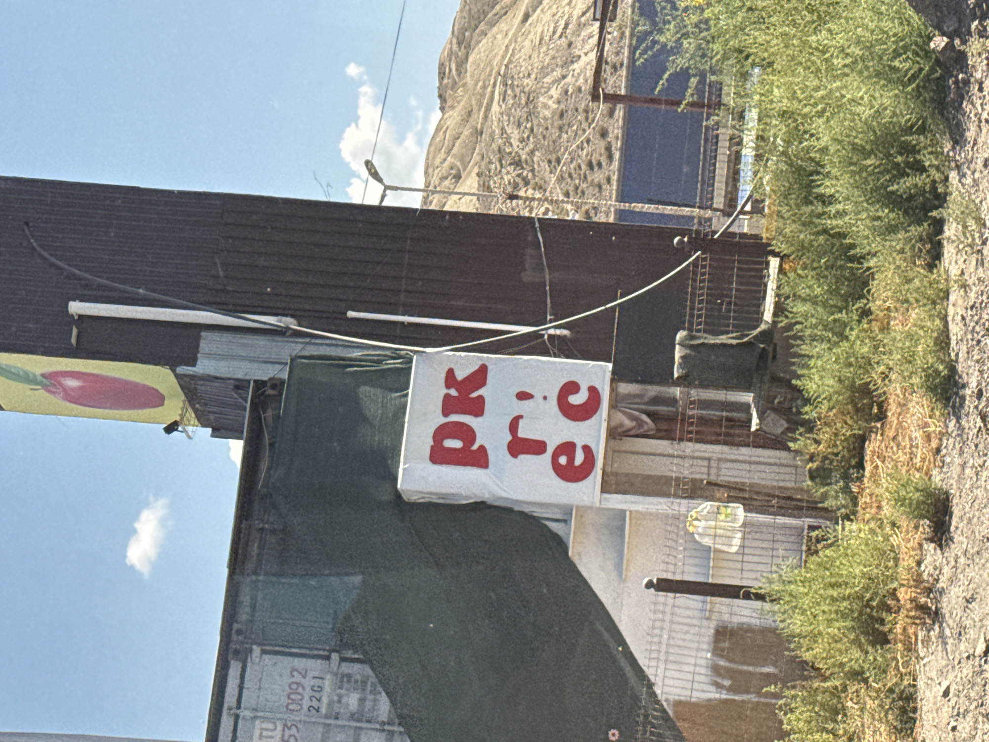 A low-angle, medium shot captures an outdoor scene dominated by a large, dark corrugated metal structure leaning to the right, against a bright blue sky with scattered white clouds. To the left of the metal structure, a large, dark green tarpaulin is draped over an object, partially obscuring it. A white sign with red lettering is attached to the front of this object. The lettering appears to read "pk" in the top line, "r!" in the middle line, and "ec" in the bottom line. Behind the tarpaulin and to the left, a white shipping container is visible, with markings "TU 53 0092 22G1" on its side. In the background, dry, barren hills rise under the sky. A streetlight with a curved arm stands near the right edge of the frame. A metal fence runs across the lower portion of the image, with some green shrubs and dry brush growing in front of and around it.