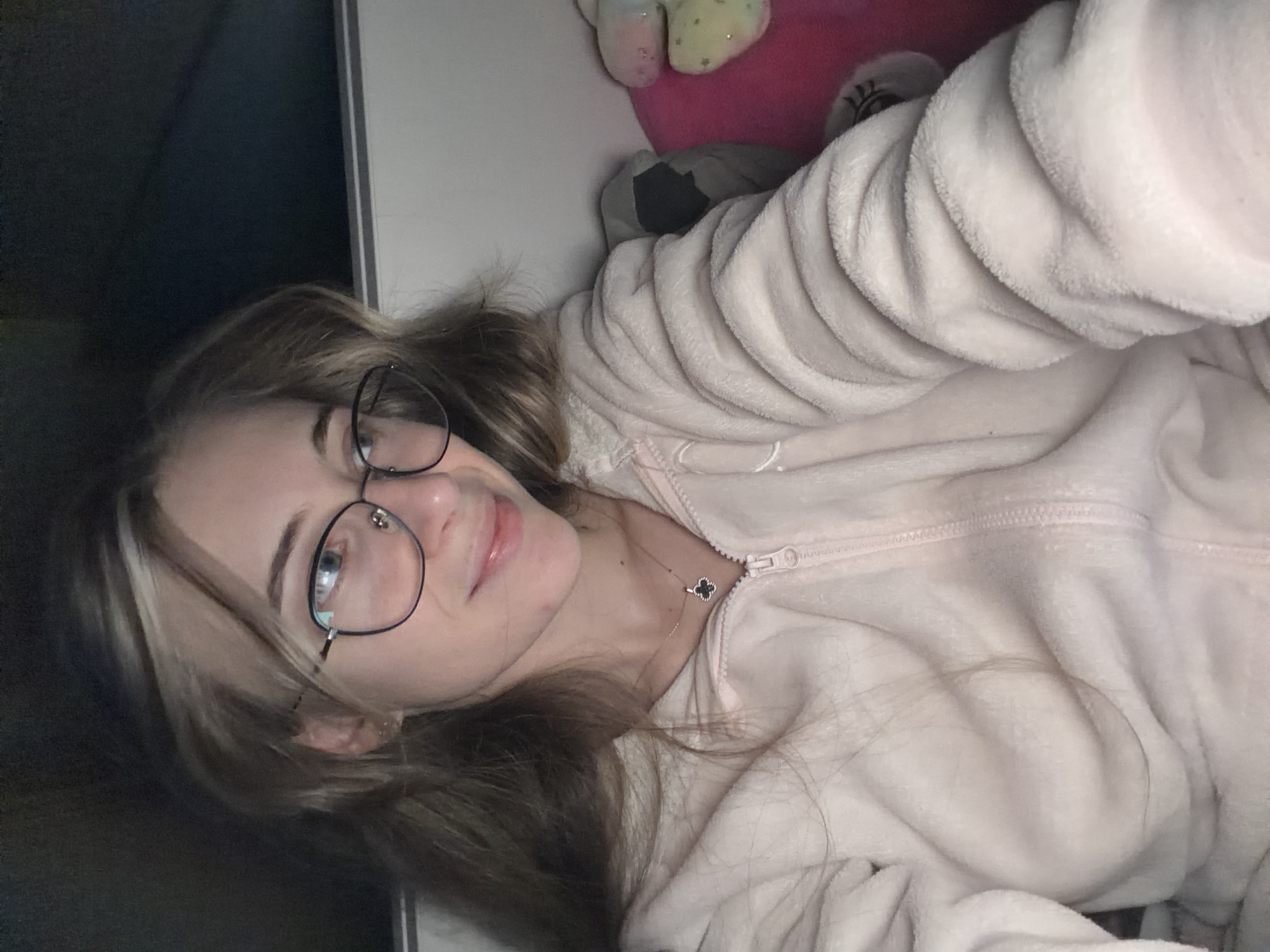 A young woman is lying on her side, wearing a light pink, fuzzy zip-up jacket and glasses. She has light brown hair with blonde streaks. She is wearing a necklace with a dark clover charm. Above her are a pink stuffed animal and a white object with yellow spots. The background includes a dark surface on the left and a white surface on the top. The location in the city of Plovdiv, Bulgaria, cannot be determined from the image.