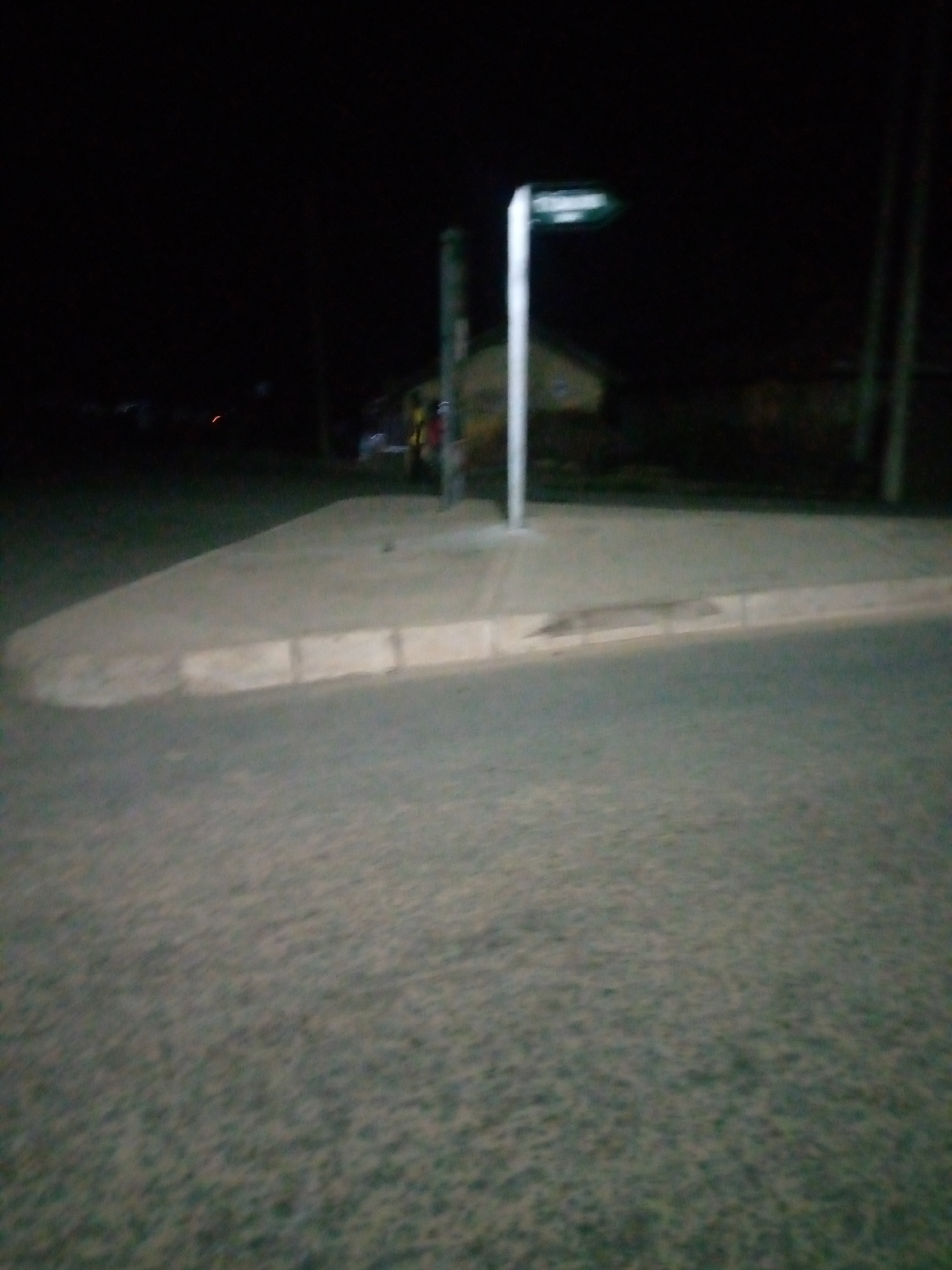 The image depicts a street scene at night in Jalingo, Nigeria. In the foreground, a paved road is visible, with a concrete curb and sidewalk to the left. The sidewalk appears to have a light-colored rectangular block pattern. In the background, a street sign with a white pole and a green directional arrow is prominently displayed. The sign has white lettering, but the text is illegible due to the image quality and lighting conditions. Behind the sign, a building with a sloping roof is faintly visible, suggesting a residential or commercial area. The overall environment is dark, with limited illumination, and the image has a slightly blurry quality. There are no discernible individuals or specific actions taking place. The scene captures a typical street corner in a Nigerian city after dark.