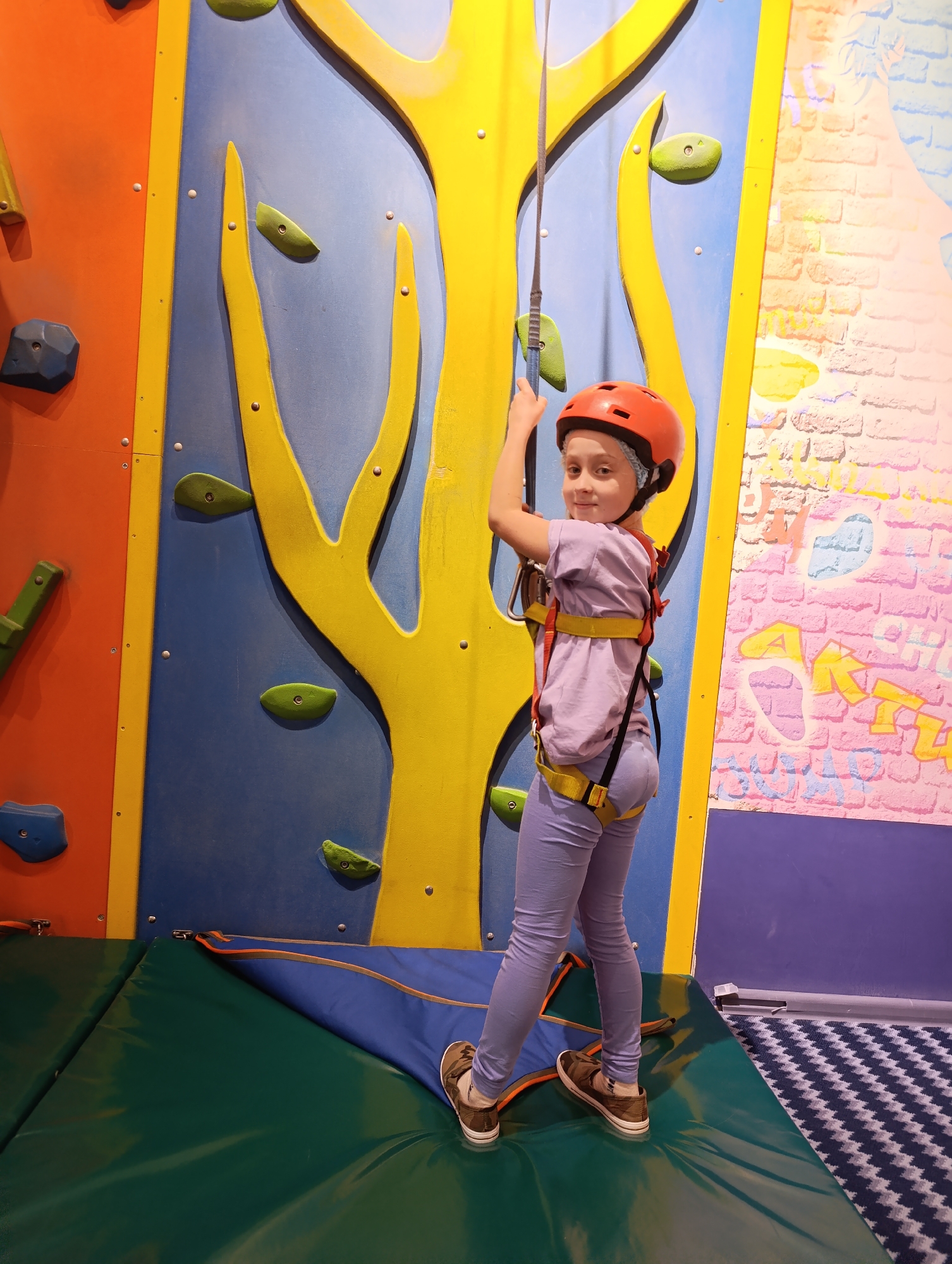 A girl wearing a helmet, harness, lilac top and pants, and brown shoes, is at an indoor climbing facility. She is attached to a rope and appears to be ready to climb a wall that features colorful panels and holds. The panels are orange, blue, and yellow. A padded mat is on the floor beneath her. The floor nearby has a blue and white zigzag patterned carpet. The city is Unknown, Unknown.