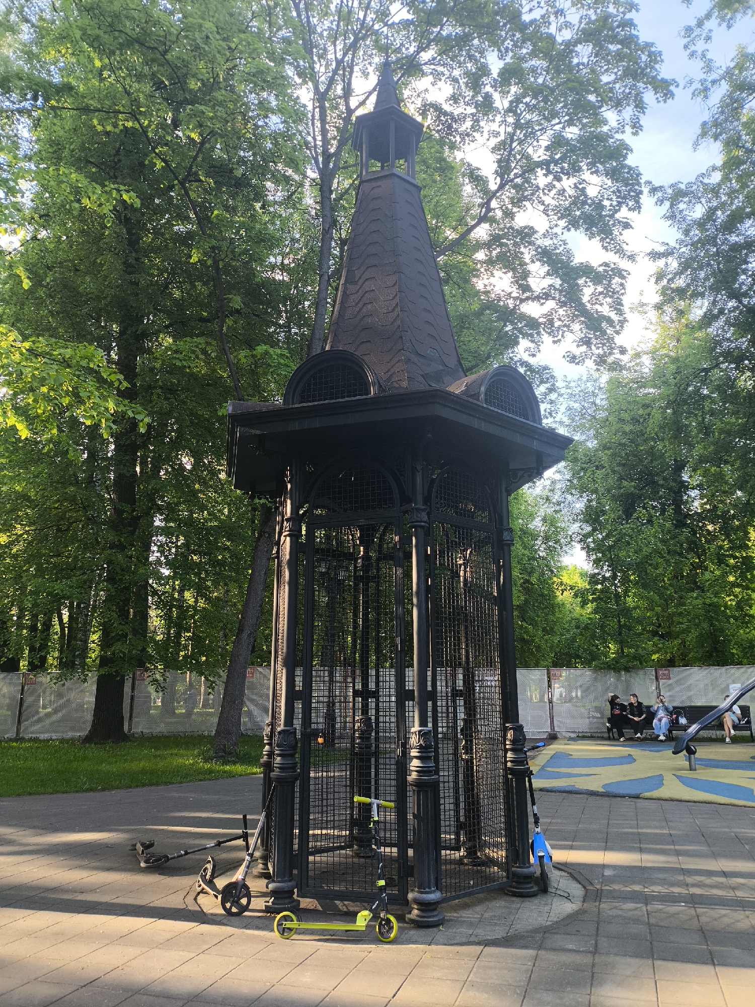 The image depicts a pavilion-like structure with a metal frame and a brown shingled roof, situated in a park. The structure has rounded windows beneath the roofline and is surrounded by scooters of various colors. A group of people is sitting on benches in the background. The pavilion is located in the Gorky Park in Moscow, Russia. The environment is characterized by lush green trees and a paved area. The lighting suggests it is daytime.