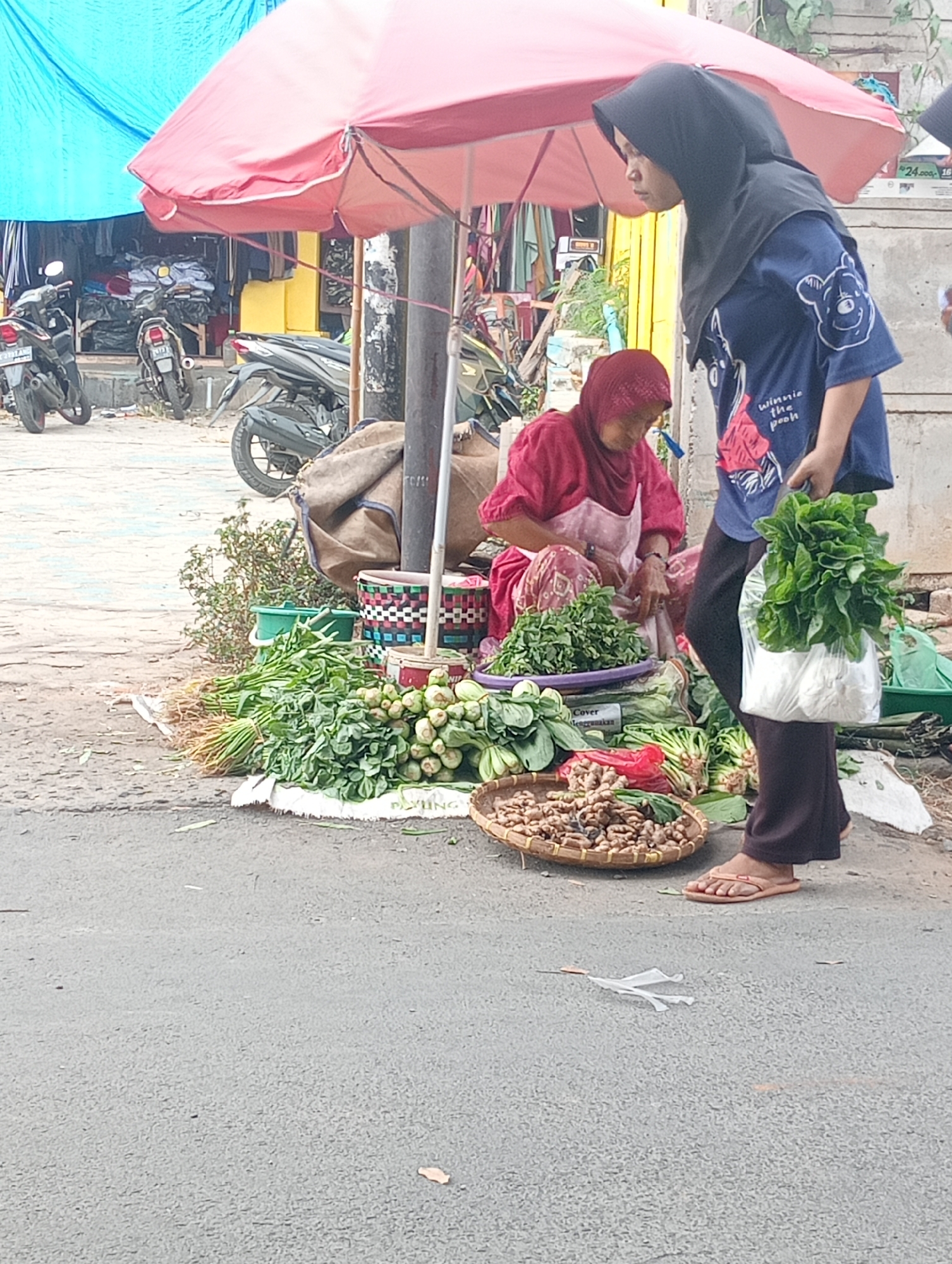 The image depicts a street scene in Bandar Lampung, Indonesia, with a focus on a woman selling vegetables from a roadside stall. The stall is set up under a large, open pink umbrella, providing shade. In the foreground, a variety of fresh produce is laid out on a white cloth, including bunches of leafy greens, what appear to be gourds or squash, and a woven basket filled with ginger or turmeric. A second woman, wearing a blue shirt with a Winnie the Pooh graphic and a black headscarf, stands to the right, holding a plastic bag filled with more greens. A third person, partially visible in a red headscarf and apron, is seated behind the vegetables, possibly preparing them. Behind the stall, there are motorcycles parked, and the blurred background suggests a marketplace or street with various shops. The ground is paved asphalt, with some debris visible. The overall impression is of a typical daily commerce scene in an Indonesian urban environment.