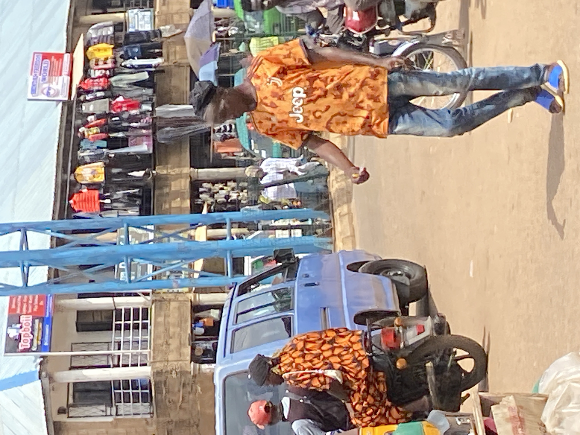 This image captures a street scene in Bukuru, Nigeria, characterized by a bustling marketplace and traffic. In the foreground, a man wearing a vibrant orange t-shirt with a "Jeep" logo and blue jeans is walking. He is accompanied by a motorcycle, suggesting he is either a rider or a passenger. Behind him, a blue van is parked. Further back, several people are visible, some standing and others seemingly engaged in market activities. Shops line the street, displaying a variety of goods, including clothing hung on racks. Blue metal scaffolding is prominent in the middle ground, likely part of a building structure or market stall. The overall environment appears to be a typical urban setting in Nigeria, with a mix of commercial activity and everyday life.