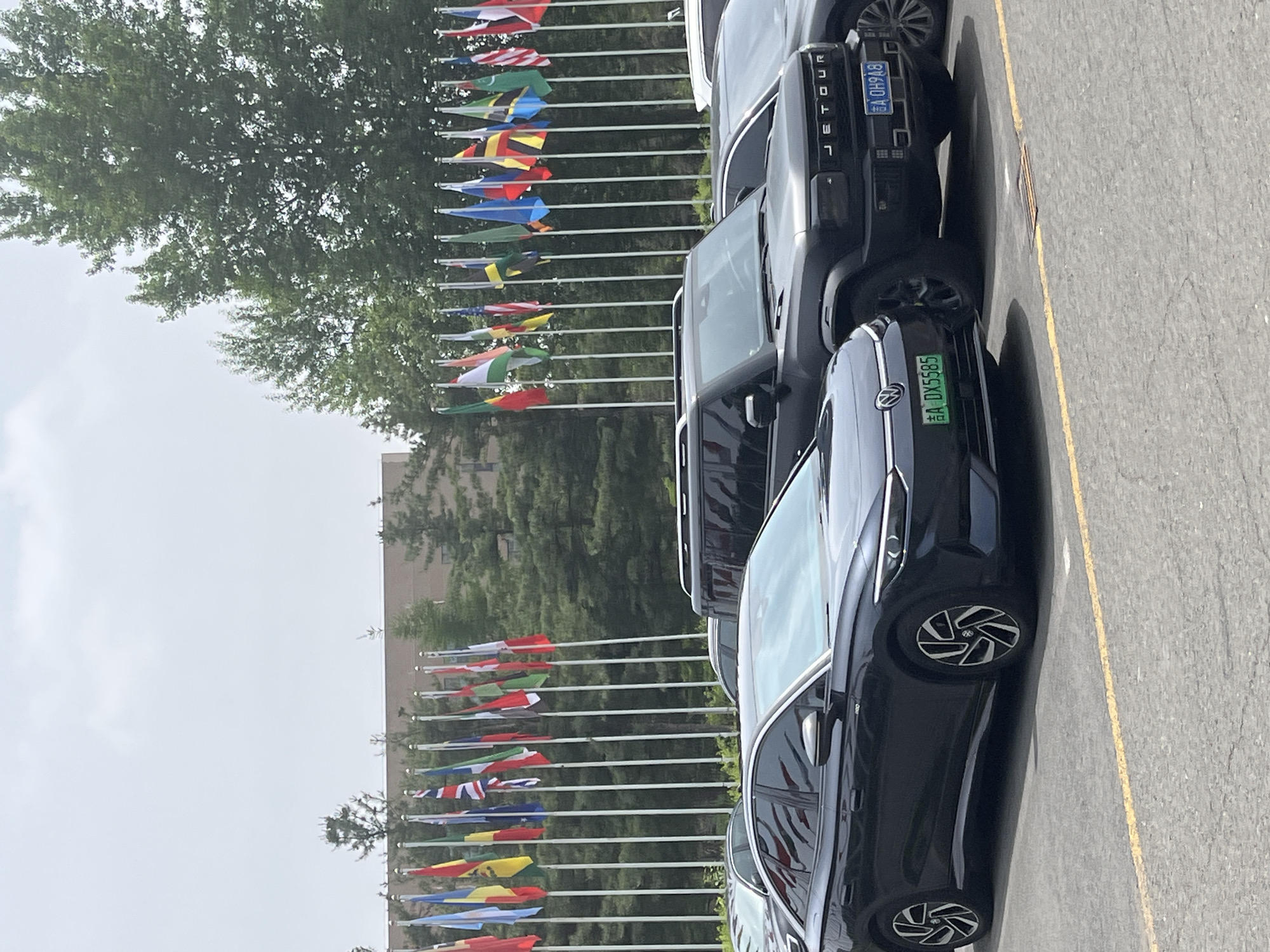 The image shows a row of cars parked along a street lined with flags. The cars appear to be in Changchun City, China, as indicated by the Chinese license plates. The flags appear to be international flags. The sky is overcast. The scene is captured during daylight hours.