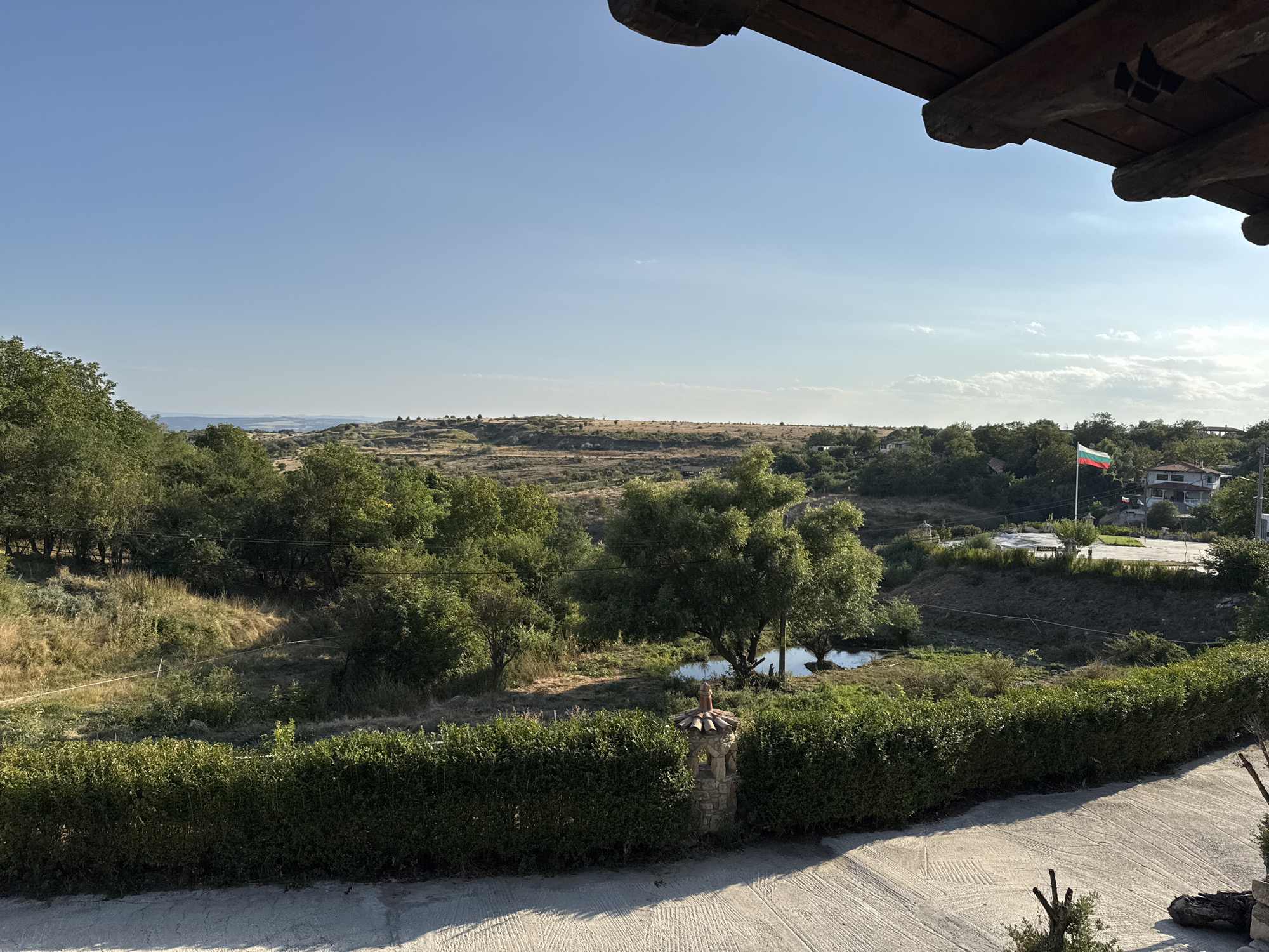 The image depicts a scenic view from a slightly elevated position, likely a balcony or porch, overlooking a rural landscape in or near Kalimantsi, Bulgaria. The foreground features a textured concrete surface with tire tracks and a small, leafless tree stump. A well-maintained, dense green hedge runs horizontally across the middle ground, partially obscuring a stone pillar topped with red roof tiles.
Beyond the hedge, the landscape unfolds with rolling hills covered in a mix of green trees and dry, light brown vegetation. A small, still body of water, possibly a pond or a very calm section of a stream, is visible nestled amongst the trees. Further in the distance, houses with white walls and red-tiled roofs are scattered amongst the greenery, indicating a residential area. A Bulgarian flag is prominently displayed on a flagpole to the right of the frame. The sky is a clear, bright blue with a few wispy white clouds. The overall impression is one of a tranquil, sunlit rural setting in Bulgaria.