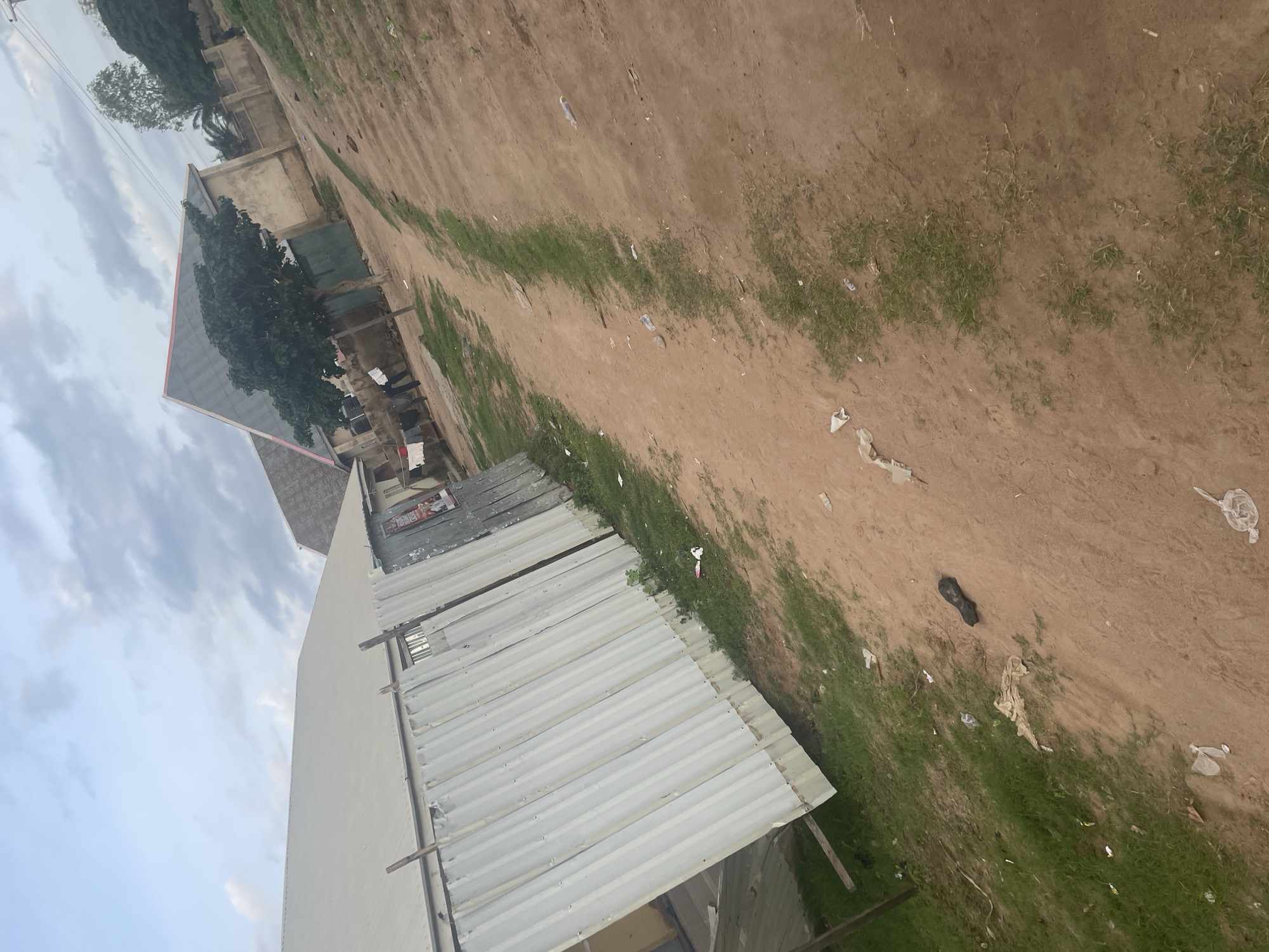 The image depicts a dirt path winding through a semi-urban environment in Jalingo, Nigeria. To the left, a corrugated metal fence forms a barrier in front of a building with a large, light-colored roof. Beyond the fence, another building with a pitched roof, possibly a house, is visible. A medium-sized tree with lush green foliage stands near this building. Several items of clothing are hung on a clothesline in front of this structure. The path itself is composed of reddish-brown soil with patches of sparse green grass growing along its edges and in the middle. Scattered across the path and the surrounding ground are various pieces of litter, including plastic bags and wrappers. In the foreground, a single dark-colored shoe lies on the dirt. The sky above is overcast with scattered clouds, suggesting it might be late afternoon or early morning. The overall impression is one of a utilitarian, somewhat unkempt, but lived-in area.