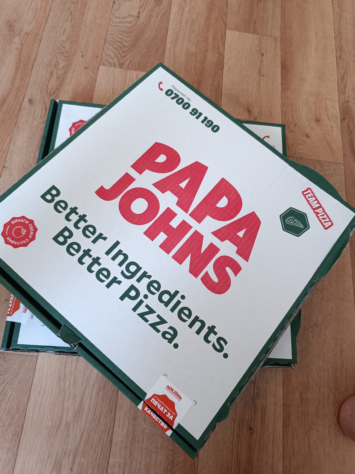 Two Papa John's pizza boxes are stacked on a wooden floor. The top box displays the Papa John's logo in red, along with the slogan "Better Ingredients. Better Pizza." Additional text includes a phone number (0700 91 190) and the phrase "TEAM PIZZA." Seals stating "Винаги Свежи Съставки" and "Печат За Качество" are visible in Bulgarian. The boxes indicate they are likely from a Papa John's in Sofia, Bulgaria, as the phone number and seals are in Bulgarian. A partial view of a foot is visible on the right.