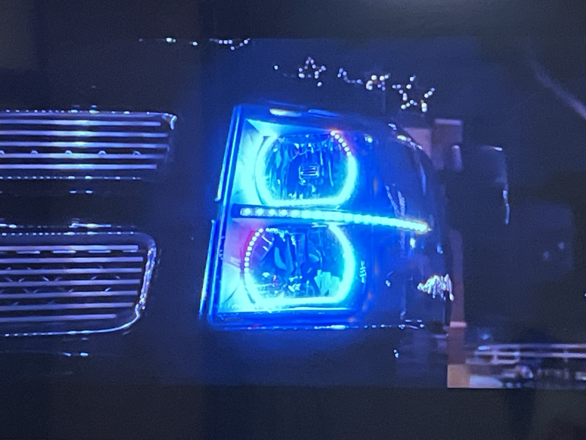 The media file shows a close-up view of a vehicle's headlights and grille. The headlights are illuminated with bright blue and red LED lights, giving them a custom, aftermarket appearance. The grille is composed of horizontal bars, also reflecting the blue light. The background is dark and blurred, suggesting it is nighttime or a low-light environment. The overall impression is of a modified vehicle, possibly a truck or SUV, with its lights on. There are scattered white lights in the upper background, resembling distant city lights or possibly decorative lights.
The setting appears to be outdoors, given the context of a vehicle. The time of day is clearly nighttime due to the darkness and the illuminated headlights being the primary light source. There are no people visible in the frame. The scene is static, focusing on the details of the vehicle's front end. The colors are dominated by the intense blue and red of the LED lights, contrasting with the dark background. No text is visible in the image.