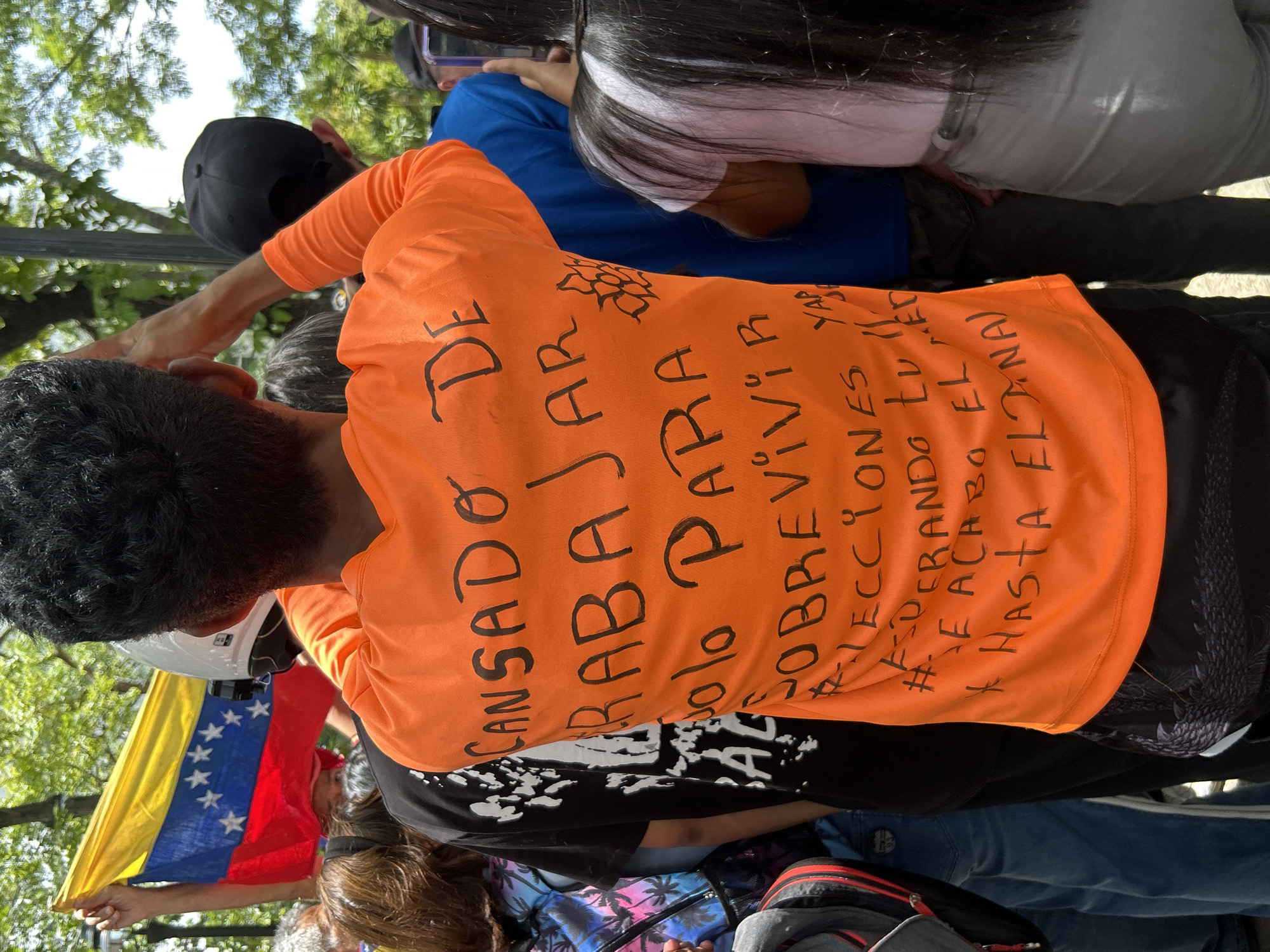 A protest or demonstration in Caracas, Venezuela is captured from an elevated perspective, showing a crowd of people gathered outdoors during daylight hours. The central focus is the back of a person wearing a vibrant orange long-sleeved t-shirt. Handwritten in black on the shirt is a clear message expressing social and political discontent. The main text reads: "CANSADO DE TRABAJAR SOLO PARA SOBREVIVIR" (Tired of working only to survive). Below this, several hashtags are visible: "#ELECCIONES YA" (#ElectionsNow), "#QUE SE ACABE EL DESGOBIERNO" (#EndTheMisgovernment), and what appears to be "+ HASTA EL FINAL" (+ Until the End). A stylized flower or sun motif is drawn above the text.
To the left of the person in orange, another individual holds a Venezuelan flag, displaying its yellow, blue, and red horizontal stripes with eight white stars in the blue band. This person is also seen with a white motorcycle helmet resting on their head. Various other participants are visible throughout the frame, some wearing hats or caps, and one person appears to be interacting with a smartphone. The setting features green tree foliage in the background against a bright sky, indicating an outdoor public space. The image conveys a sense of public frustration and a call for political change in Venezuela.