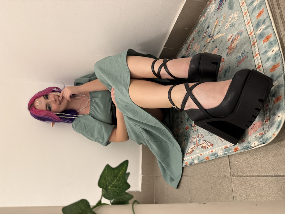A young woman with purple and pink hair, pointed ears, and a silver headpiece smiles, wearing a light green dress and black platform shoes with crisscrossing straps. She leans against a white wall near a houseplant, and a patterned rug is visible beneath her feet. The overall lighting is soft and natural.