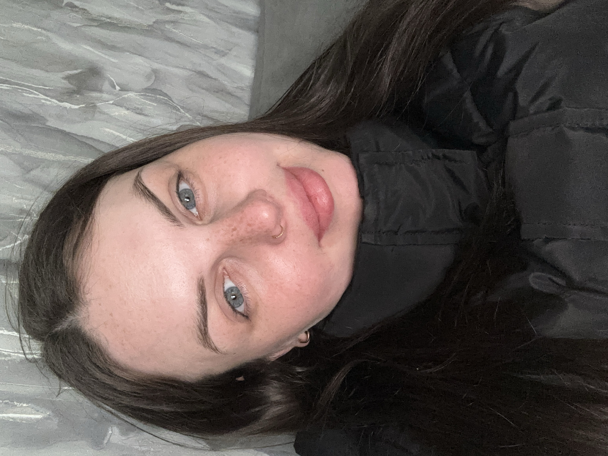 This is a portrait of a young adult female lying down, looking directly at the camera. She has long, dark brown hair, light blue eyes, and fair skin with noticeable freckles across her nose and cheeks. She wears a small golden hoop nose ring on her left nostril and a small golden hoop earring in her right ear. Her lips are full and have a natural pink hue. She is dressed in a dark, possibly black, quilted or puffy jacket with a prominent collar.
The background features a light grey surface with a subtle, swirling marble pattern, suggesting a bedspread, blanket, or upholstered surface. The image appears to be taken indoors, under soft, even lighting, with no indication of natural light. The subject's expression is neutral and relaxed, conveying a sense of calm. No text is visible within the image. The scene is set in what appears to be an indoor location in Chornomorskoe, Ukraine.