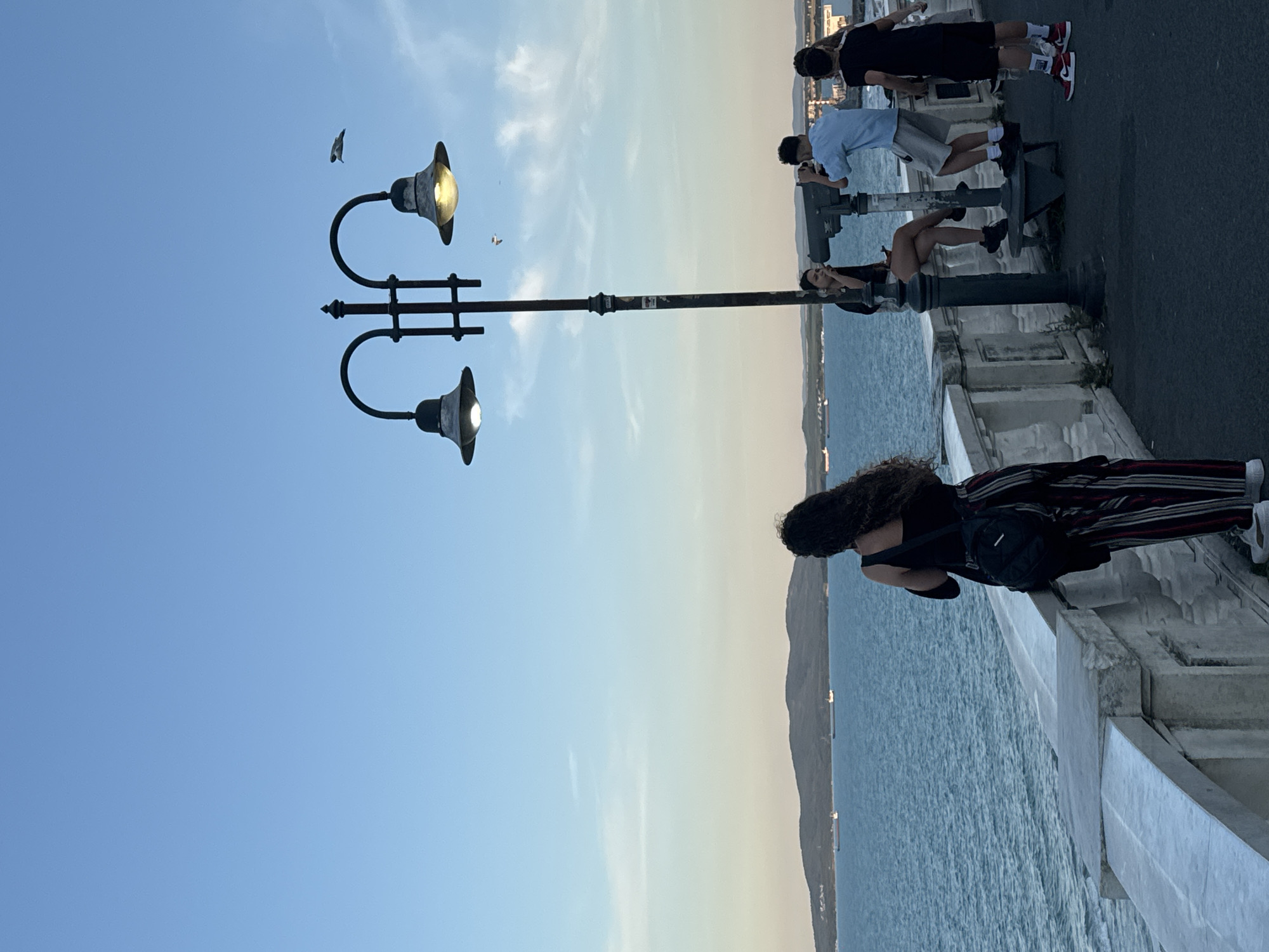 The image shows a waterfront promenade in the city of Burgas, Bulgaria. The sea is to the left, with a distant coastline featuring hills. Several cargo ships are visible on the water. In the foreground, a woman with long curly dark hair, wearing a black top and striped trousers, stands facing the sea. To her right, a lamppost with two lights stands near a pair of coin-operated binoculars. A woman sits on a bench by the binoculars, and two men stand nearby. One man, wearing a light blue shirt and shorts, is looking through the binoculars. The other man, wearing a black t-shirt and shorts, stands to his right. The sky is a clear blue with a few wispy clouds. The overall lighting suggests it is late afternoon or early evening.
