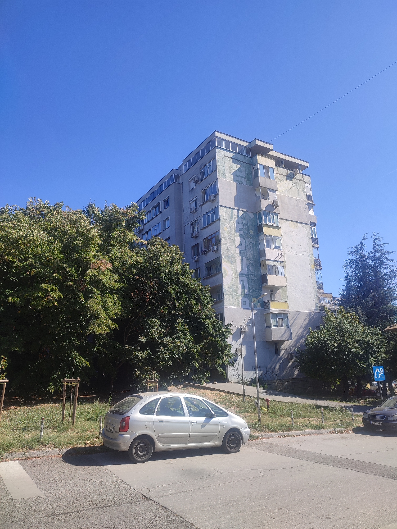 In Varna, Bulgaria, on a clear, sunny day, a multi-story apartment building with a faded mural on its facade stands tall. A silver hatchback car is parked on the street in front of the building, casting a shadow. Adjacent to the car, a pedestrian crossing is marked on the asphalt. Lush green trees line the street, some partially obscuring the building. In the background, another car is visible, and a blue sign indicating pedestrian crossing is mounted on a pole. The environment appears to be a typical urban residential area.