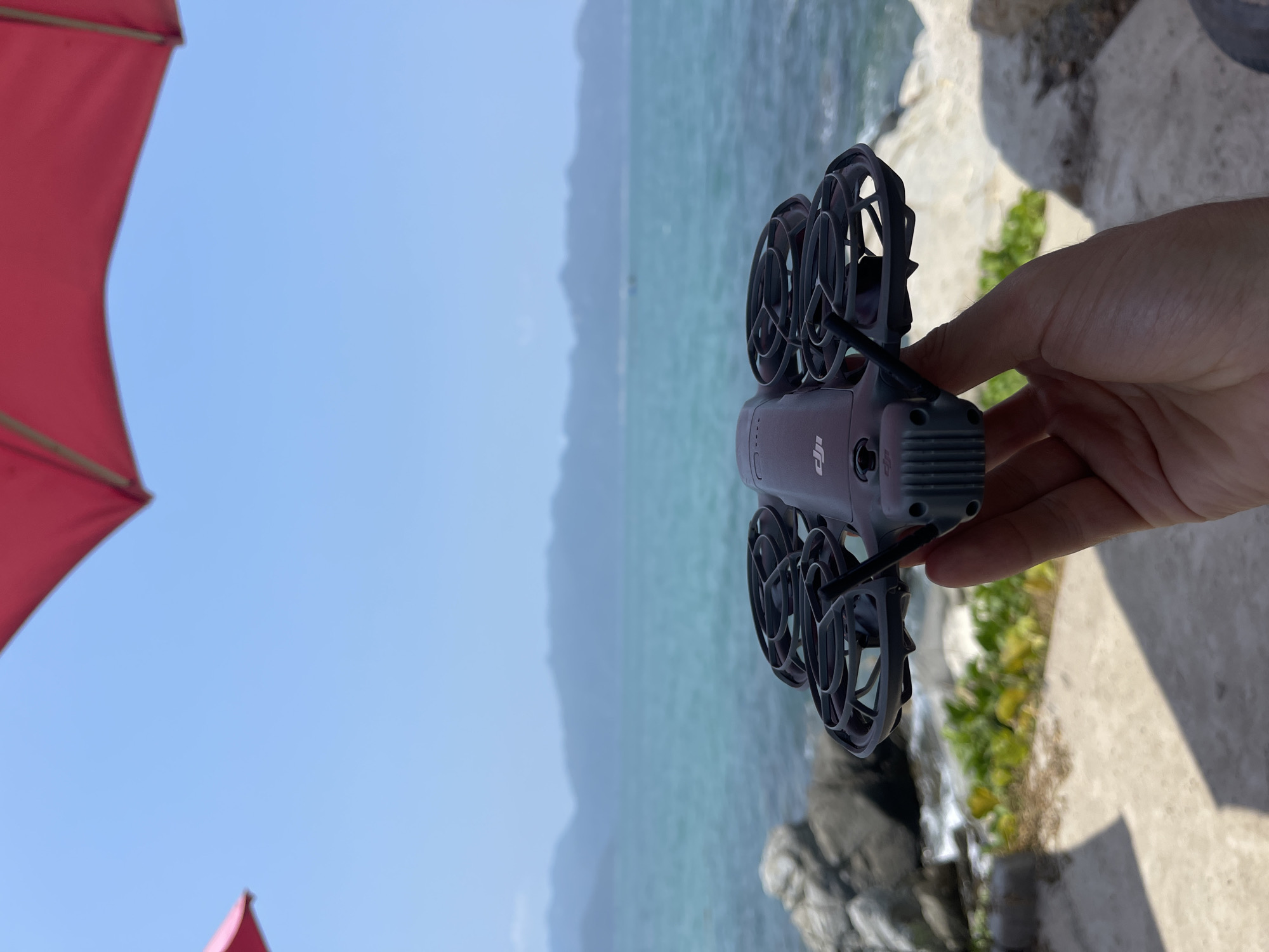 A handheld DJI drone is oriented vertically in the foreground of the photograph. The drone is gray and has circular propeller guards. A hand is holding the drone. The background consists of an ocean scene with visible hills in the distance under a clear sky. Parts of a red canopy are visible in the upper left and lower left corners. Rocks and some greenery are in the lower right corner. The location is Vĩnh Lương, Vietnam.
