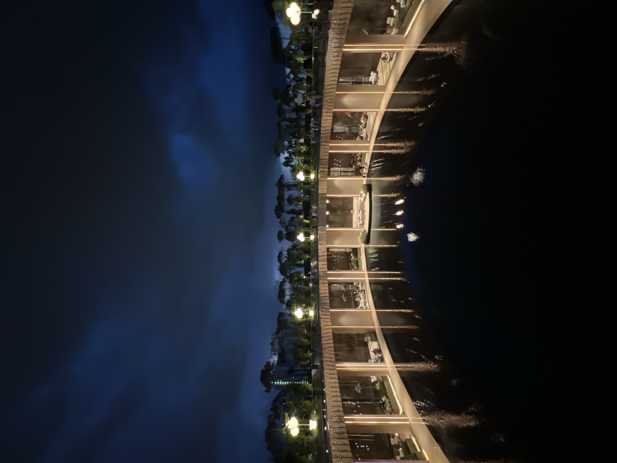 The image shows a dimly lit, open-air structure at night, featuring a semi-circular design with a water feature in the foreground. The structure has a textured, possibly stone or metal, facade with large glass windows that reveal interior seating areas. Vertical lights illuminate the facade, casting reflections on the dark water. A line of trees with distinctive spherical lamps stands on the upper level of the structure, against a dark, cloudy sky. Some people are visible seated or walking within the interior spaces, and near the water's edge. The overall scene suggests an outdoor dining or lounge area, possibly part of a resort or park. The location is identified as Krasnodar, Russia.