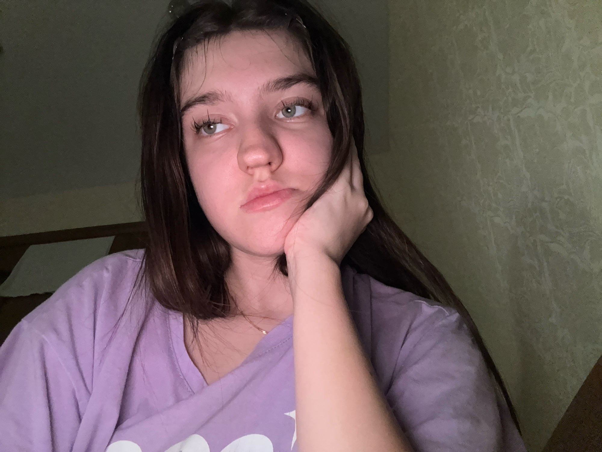 A young woman with long, dark brown hair and light green eyes is captured in a close-up, self-taken portrait, likely in a domestic setting in Zavolzhye, Russia. She is wearing a light purple t-shirt. Her right hand is propping up her face, resting on her cheek and jawline, and she is looking away from the camera with a slightly bored or thoughtful expression, her lips slightly parted. The background appears to be an interior wall with a textured pattern, dimly lit by an unseen light source, casting shadows. To the left of the frame, a portion of a dark wooden structure and a white object, possibly bedding, are visible.