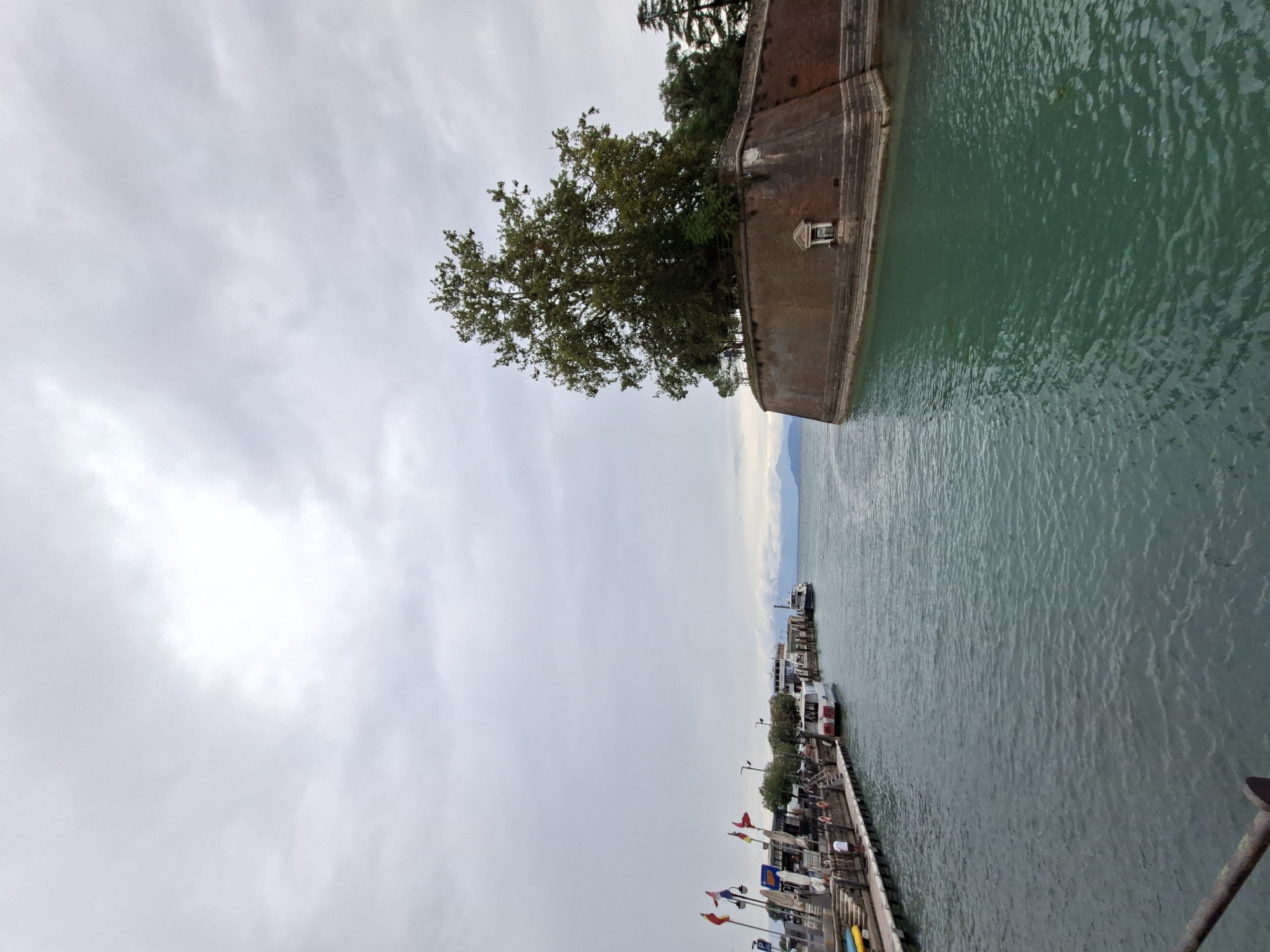 The image captures a view of the harbor in Peschiera del Garda, Italy, with a prominent brick fortress on the right and a dock with several boats on the left. The sky is overcast, creating a diffused light. The water in the harbor is a greenish-blue color. On the left, the dock is lined with boats of various sizes, and several people are visible walking and standing on the dock. Flags are flying from poles on the dock. The fortress, constructed from reddish-brown bricks, has a large tree growing on top. A small, decorative window is visible on the face of the fortress. In the distance, mountains can be seen through the hazy sky.