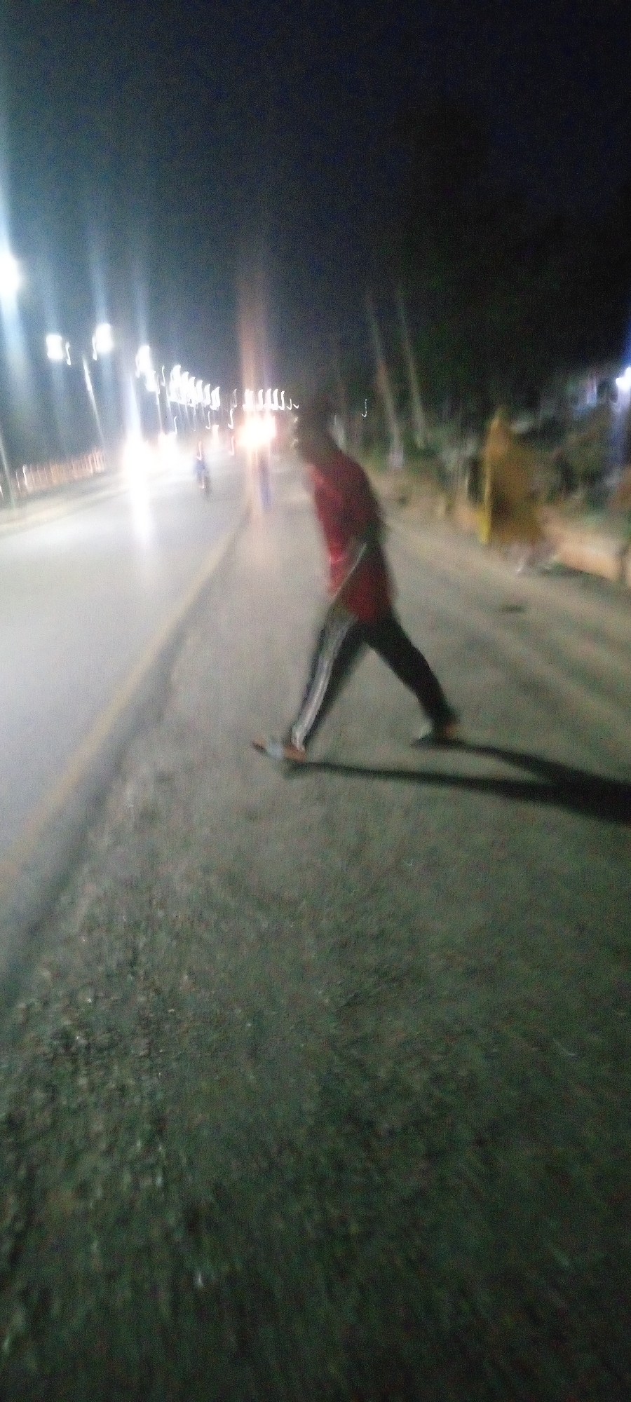 At night in what appears to be Zango-ndaji, Nigeria, a person wearing a red shirt and dark pants walks along the side of a road, possibly near its median. The person's features are blurred. Street lights are visible, along with the headlights of oncoming traffic in the distance on the left side of the frame. A motorcyclist is also visible. A figure in yellow is also blurred, possibly also a pedestrian on the side of the road. The road surface appears to be dark and wet. The overall image quality is low, with motion blur and limited detail.