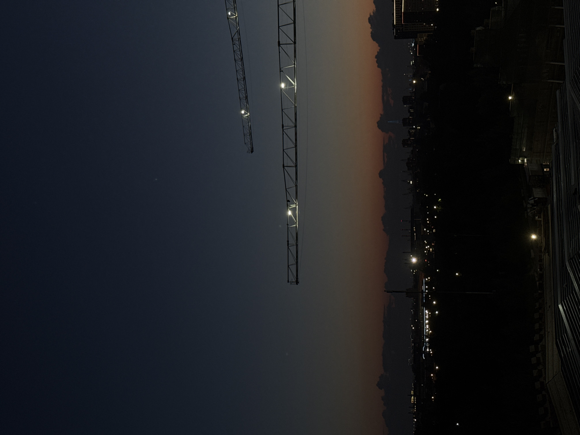 The image depicts an aerial view of a city at dusk. Two construction cranes, illuminated by spotlights, dominate the upper portion of the frame, stretching across the dark blue sky. Below them, a silhouetted cityscape unfolds, characterized by a scattering of lights from buildings and streetlamps. A thin band of orange and red hues at the horizon suggests the last remnants of sunset. The foreground is partially obscured by dark, indistinct structures, possibly other construction sites or rooftops. The overall atmosphere is one of urban development against a fading sky. The location is identified as the city of Sinyavino, Russia.