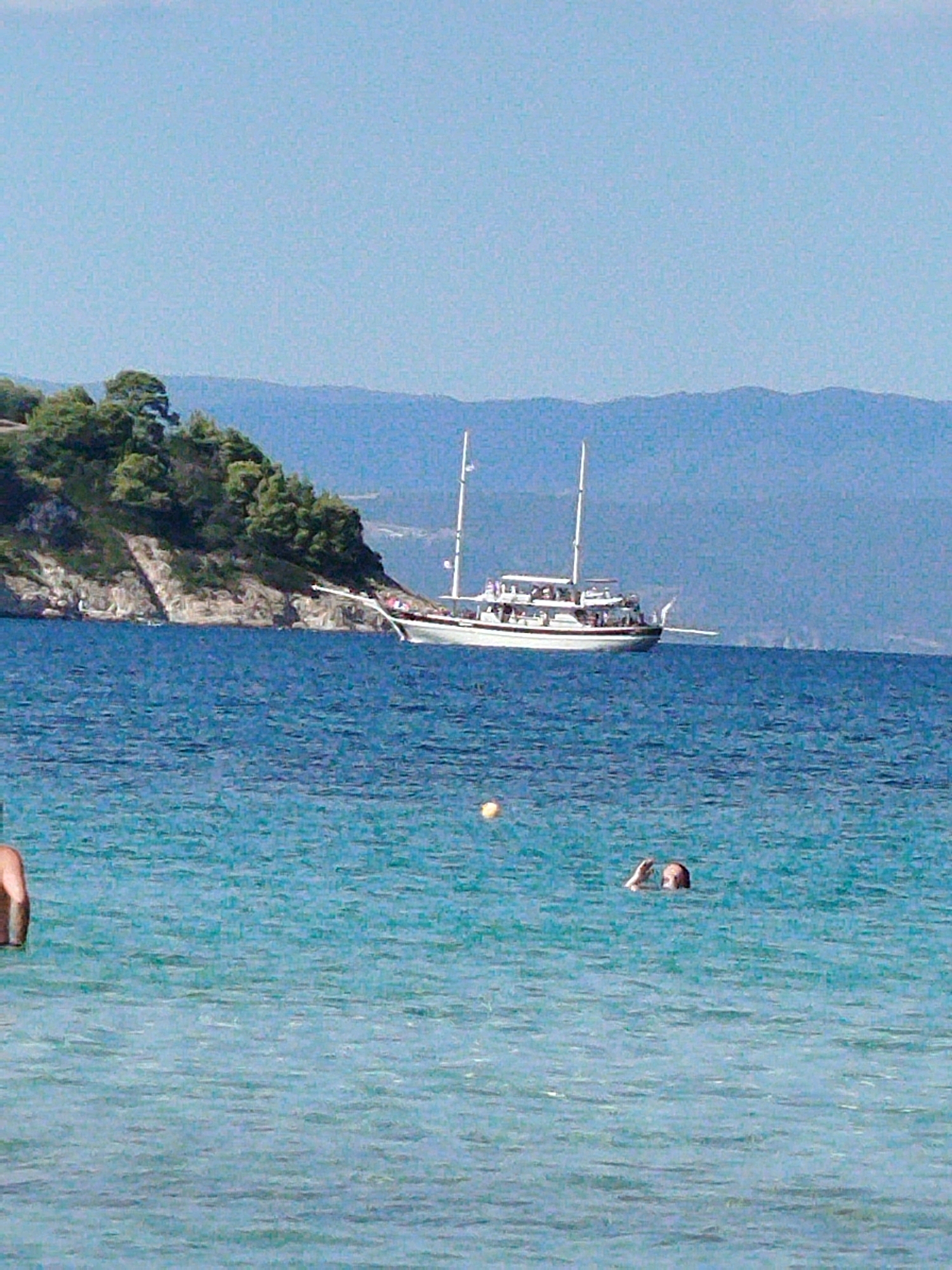 This is a medium-shot, horizontal, eye-level photograph taken on a sunny day in the Sithonia Municipal Unit, Greece. The image depicts a vibrant blue sea in the foreground, with gentle waves rippling on the surface. A person's left arm is visible on the left edge of the frame, partially submerged in the water. Further into the water, a person's head and raised right hand are visible above the surface, appearing to swim or wave. In the mid-ground, a large, white sailing boat with two masts is anchored or moored in the sea. The boat has multiple decks and passengers visible on its upper levels. Behind the boat, a lush, green, rocky hillside is visible, dotted with trees. In the far background, hazy blue mountains form the horizon under a clear, light blue sky. A small, yellow buoy is visible in the water to the left of the sailing boat. The overall scene conveys a sense of leisure and vacation.