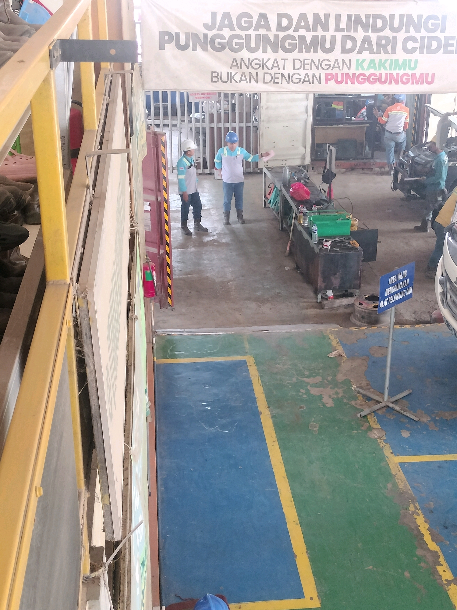 The image depicts an interior workshop or garage setting. In the foreground, a yellow metal railing and yellow vertical support beam are visible, partially obscuring the scene. A large white banner with Indonesian text hangs across the upper portion of the image. The text on the banner reads "JAGA DAN LINDUNGI PUNGGUNGmu DARI CIDERA" and "ANGKAT DENGAN KAKIMU BUKAN DENGAN PUNGGUNGmu," which translates to "Protect and protect your back from injury" and "Lift with your legs, not with your back."
In the midground, several individuals in work attire are present. One person, wearing a light blue shirt and dark pants, stands near a workbench. Another individual in an orange and grey work shirt and blue jeans is seen near a vehicle. A third person in a green and grey uniform is working on a vehicle. The workshop contains various tools, equipment, and a car. A blue sign with Indonesian text stating "AREA WAJIB MENGGUNAKAN ALAT PELINDUNG DIRI" (Mandatory area to use personal protective equipment) is prominently placed on the floor.
The floor of the workshop is a mix of concrete and painted surfaces, with distinct blue and green sections marked by yellow lines. Various items, including bottles, a green toolbox, and what appears to be a spare tire, are scattered around the work area. The environment appears to be functional and utilitarian, characteristic of a maintenance or repair facility. The image does not contain specific visual cues to pinpoint its exact location within Maburai, Indonesia, beyond it being an industrial or workshop environment.