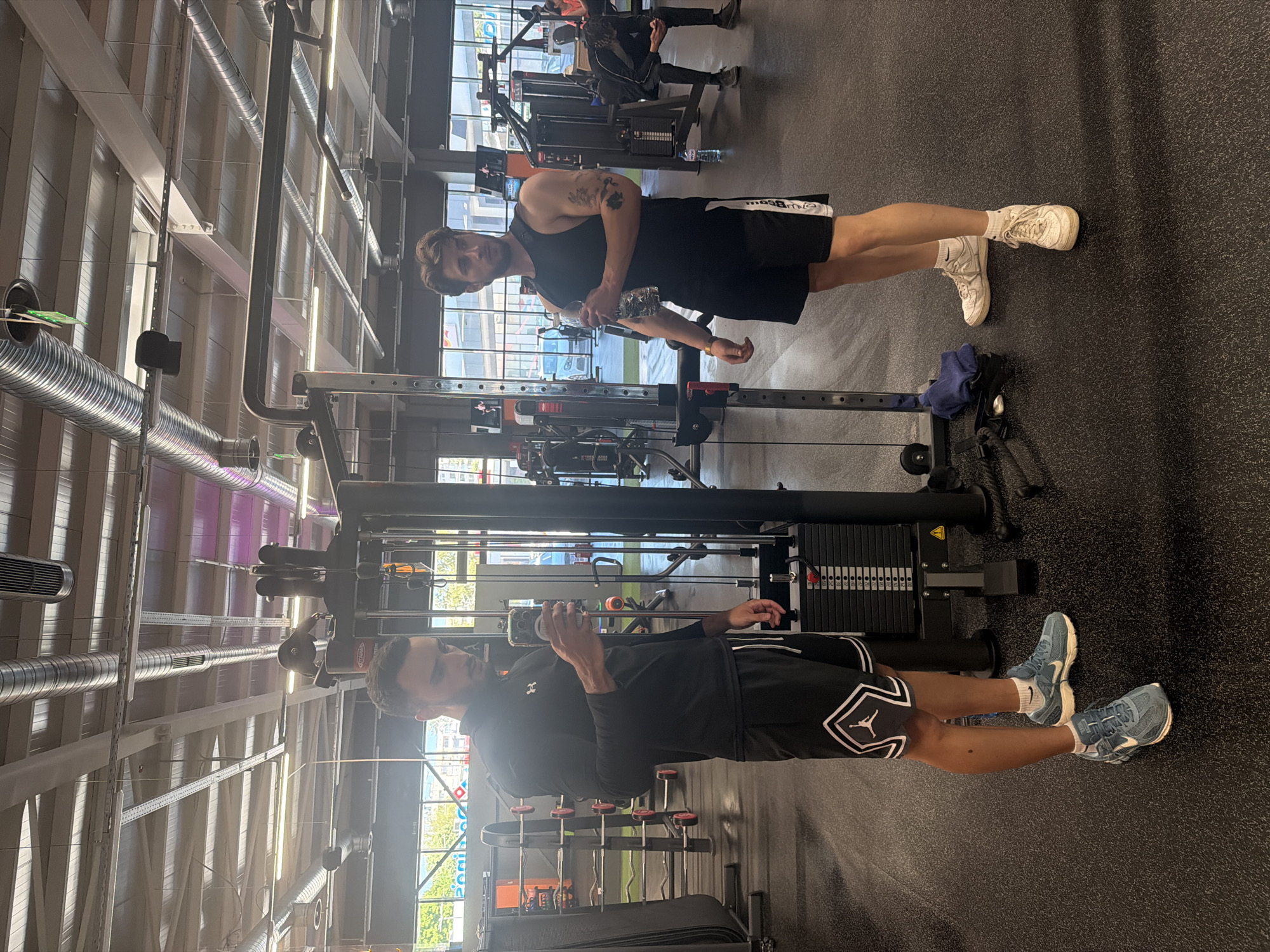 Two men are in a gym. One man is wearing a black tank top and black shorts with white sneakers. The second man is wearing a black long sleeve shirt and black shorts with white accents, and blue sneakers. The men are both holding water bottles near a weight machine. The gym has dark flooring and exposed ductwork on the ceiling. Outside the window are green trees and buildings. The location is a gym, but cannot be determined to be in Plovdiv, Bulgaria from the image.